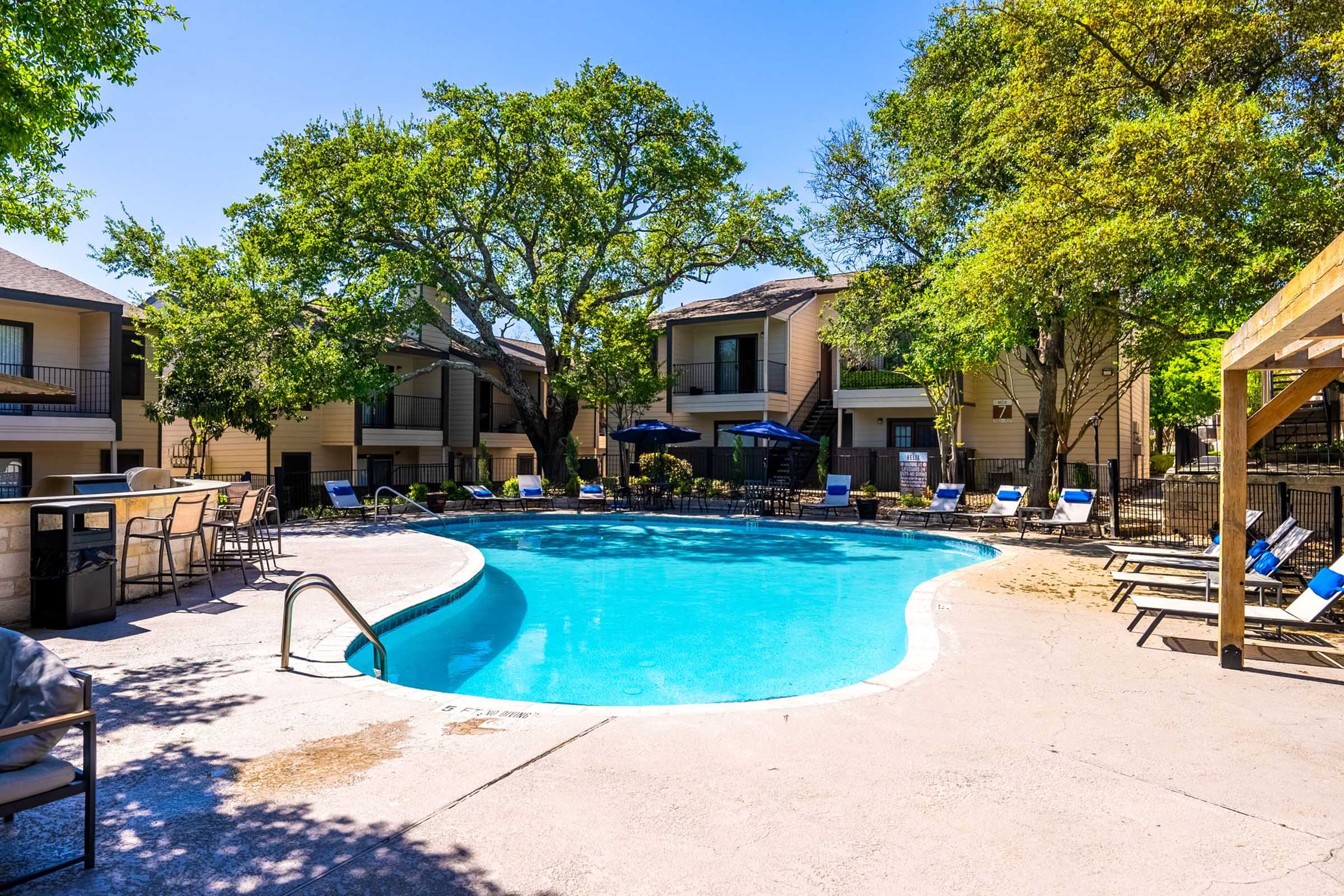 A sunny outdoor swimming pool surrounded by lounge chairs and small tables. The pool is situated among residential buildings with greenery, including large trees, providing shade. Umbrellas are set up for added comfort, creating a relaxing atmosphere.