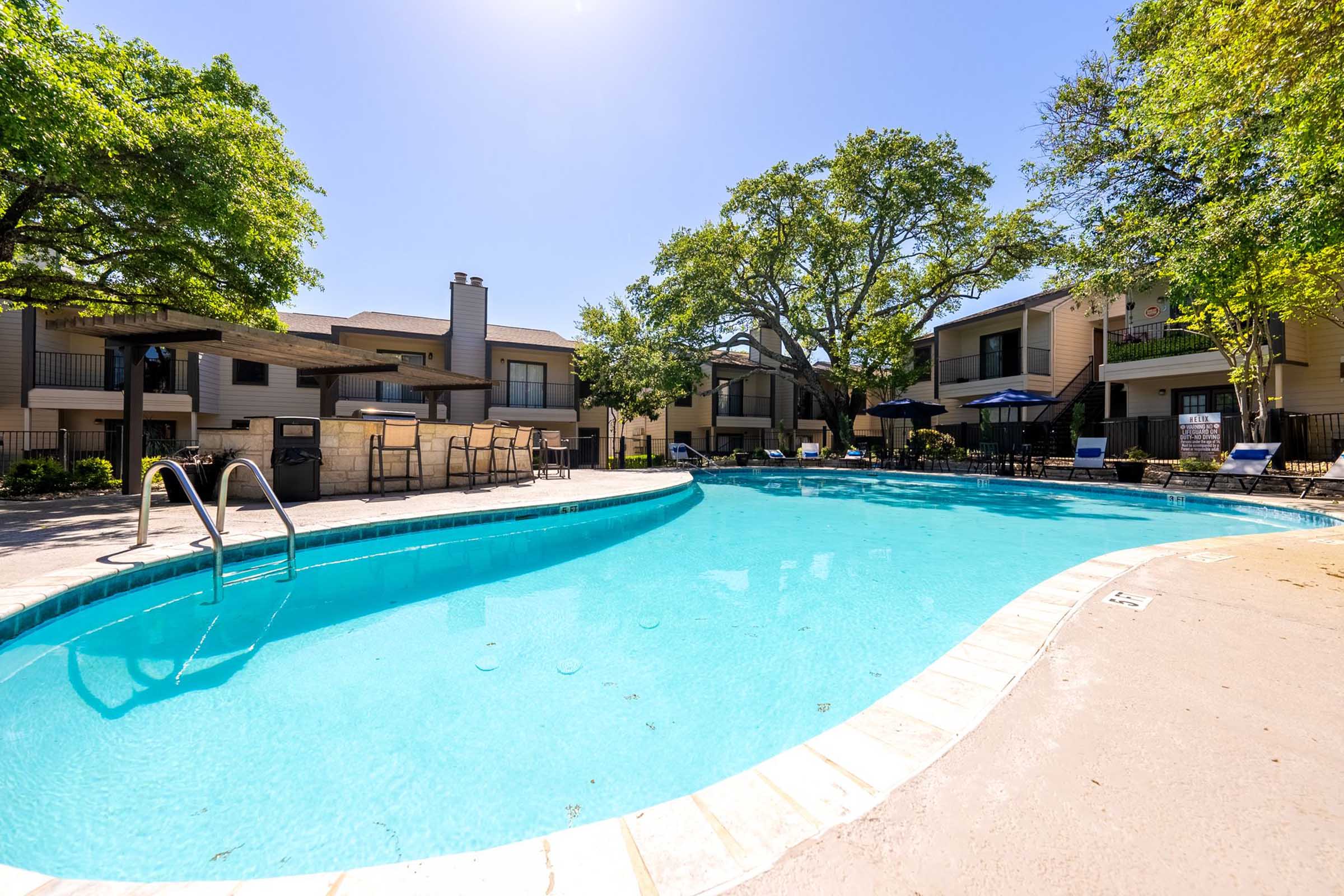 Bright, inviting outdoor swimming pool surrounded by lounge chairs and shaded tables. Lush green trees provide a pleasant backdrop, and modern apartment buildings are visible in the background under a clear blue sky. The area is ideal for relaxation and leisure.