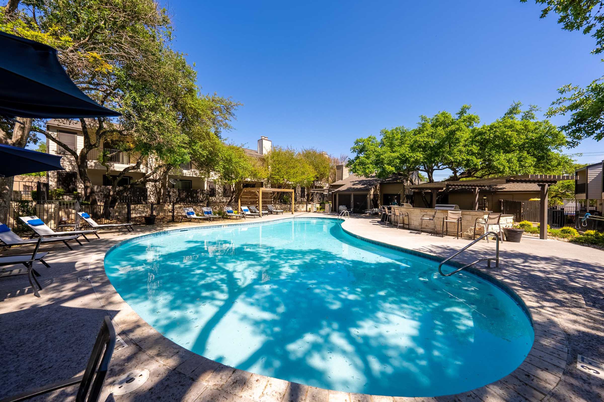 A clear blue swimming pool surrounded by lounge chairs and umbrellas, with lush green trees and buildings in the background under a bright blue sky. The peaceful setting invites relaxation and leisure.