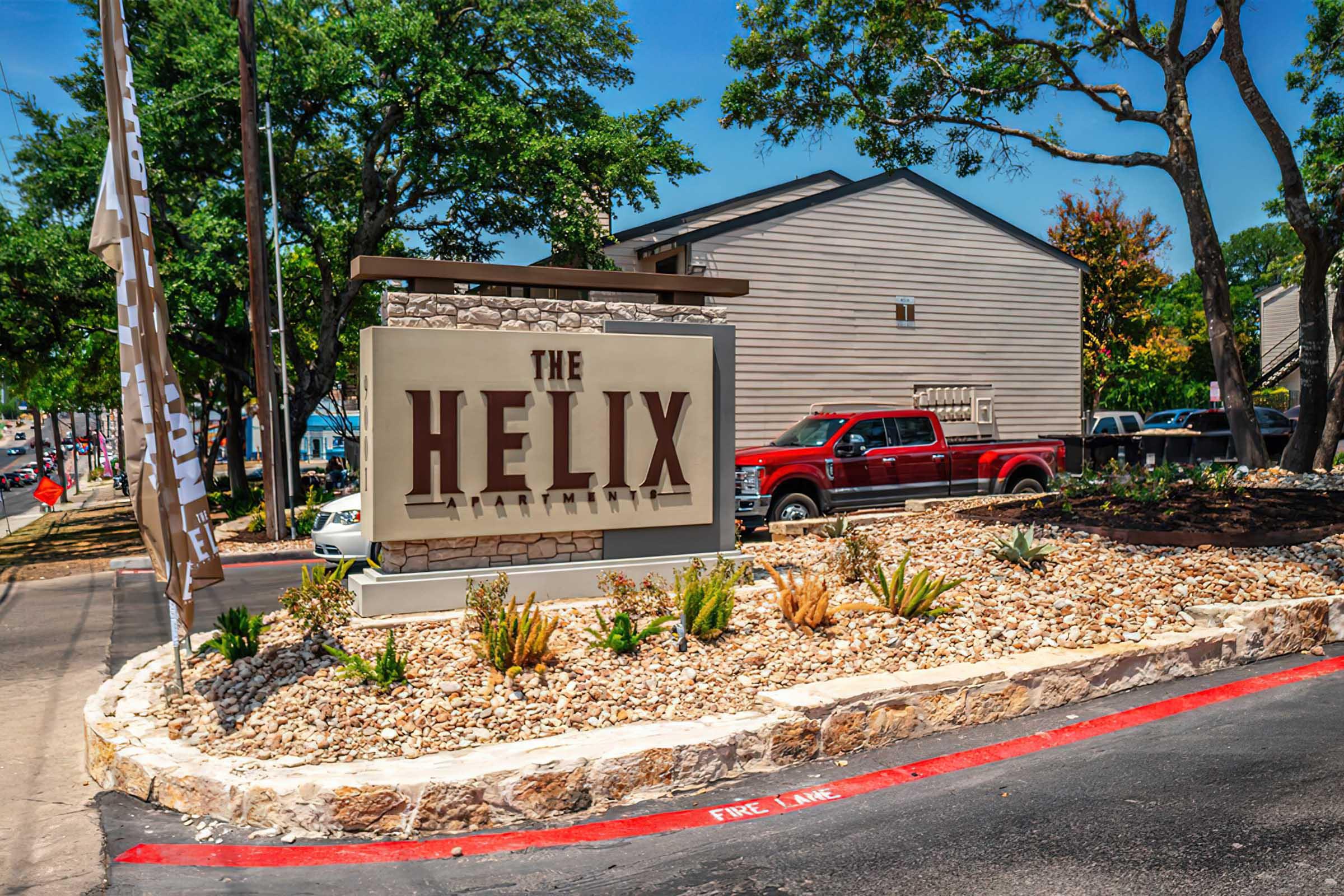 A sign for "The Helix Apartments" surrounded by landscaping, featuring rocks and small plants. A parking area is visible, with a red pickup truck parked nearby. Trees line the background under a clear blue sky.