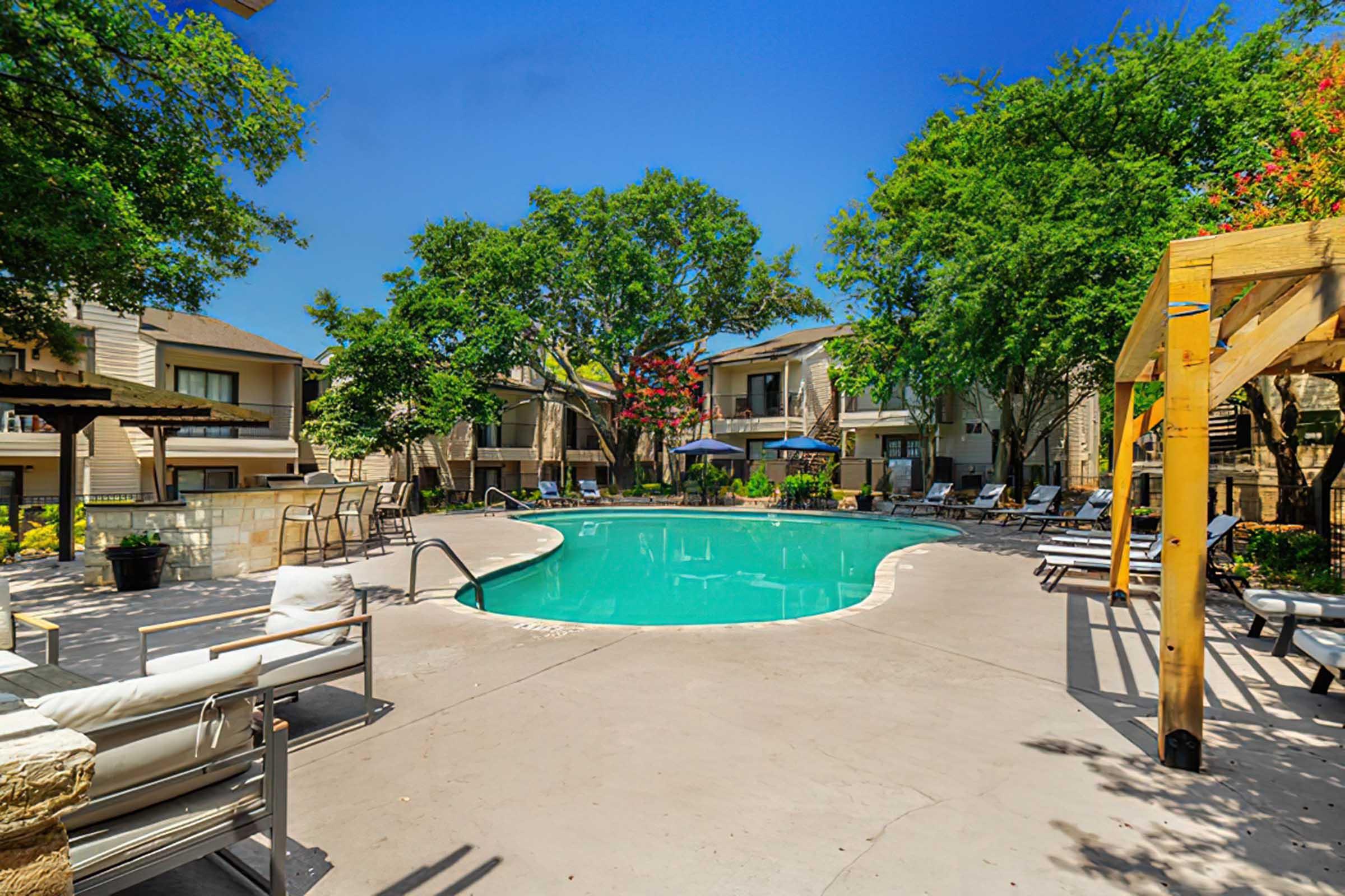 A clear swimming pool surrounded by lounge chairs and shaded areas. Green trees and colorful landscaping enhance the outdoor space. Nearby, there are several umbrellas and a pergola, creating a relaxing atmosphere in a residential complex. Blue sky above adds to the inviting scene.