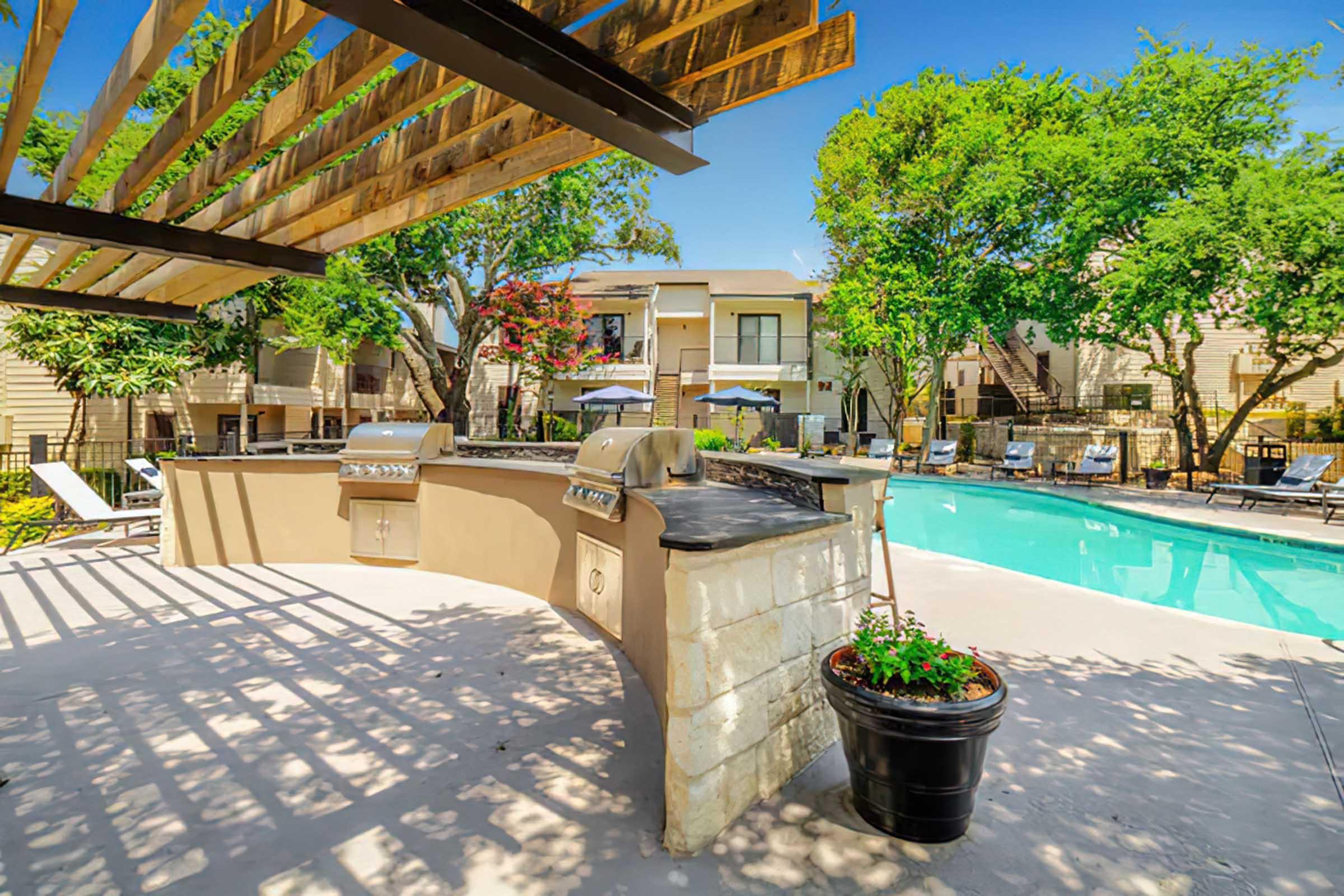 A scenic outdoor area featuring a swimming pool, surrounded by lounge chairs and greenery. There is a covered grilling station with two BBQ grills and a potted plant, creating a perfect space for relaxation and outdoor cooking. The background includes residential buildings and vibrant trees under a clear blue sky.