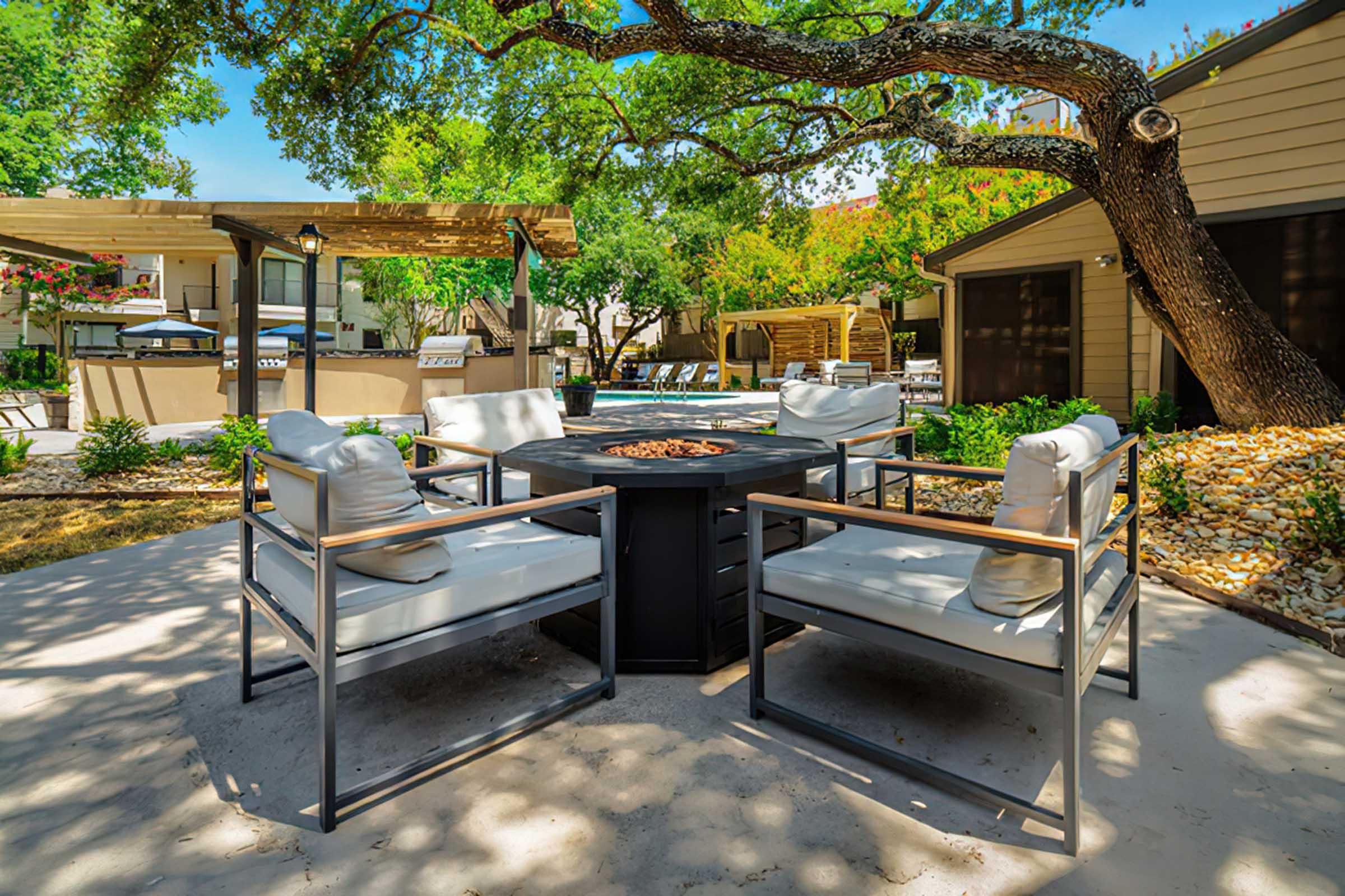 A cozy outdoor seating area featuring four white cushioned chairs arranged around a black fire pit, surrounded by well-maintained greenery and trees. In the background, there are shaded structures and a glimpse of a swimming pool, creating a tranquil and inviting atmosphere.