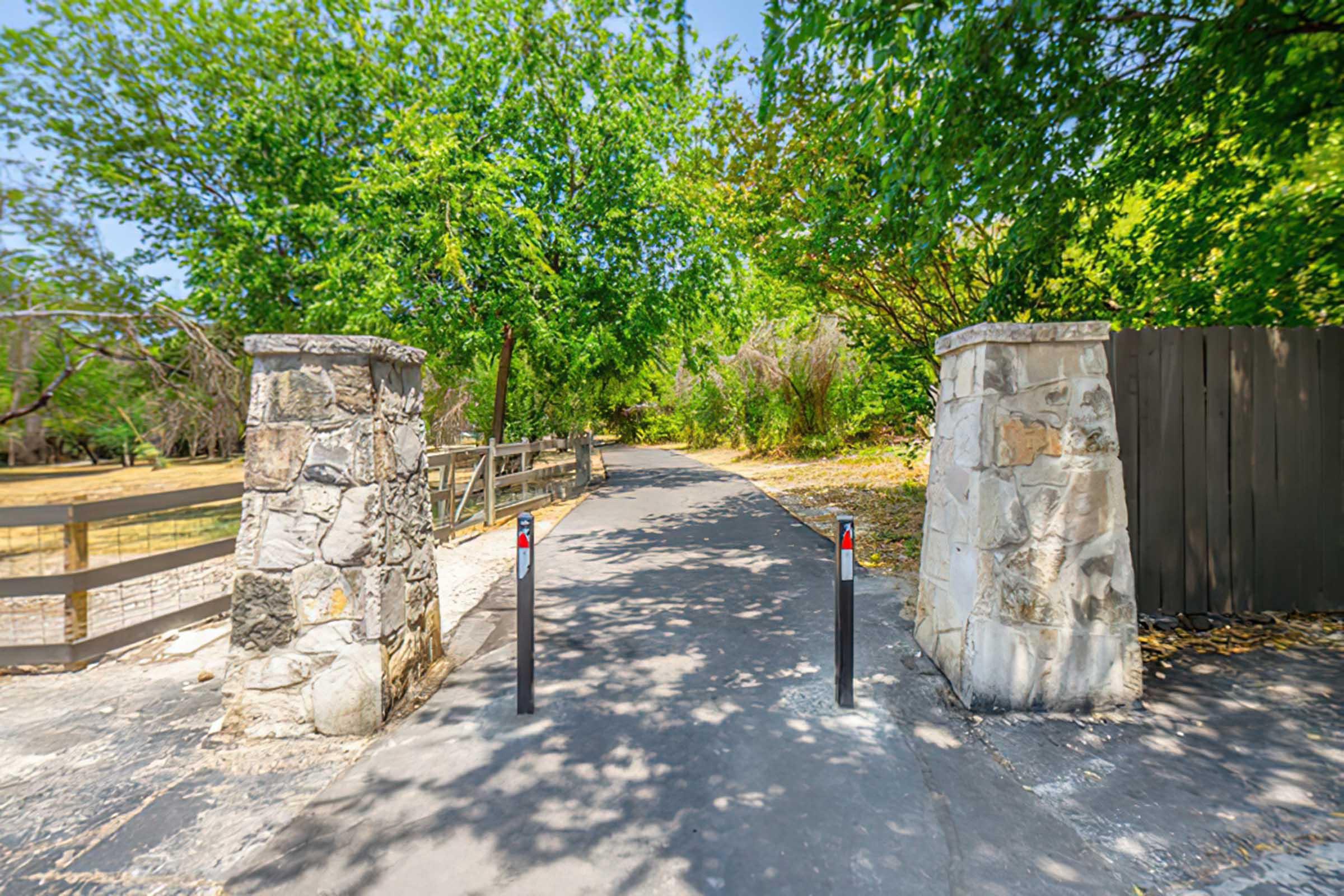 A scenic pathway lined with greenery, leading through stone pillars that mark the entrance to a park or nature area. The pathway is paved and flanked by trees, creating a peaceful and inviting atmosphere. A wooden fence runs alongside the path, enhancing the natural setting.