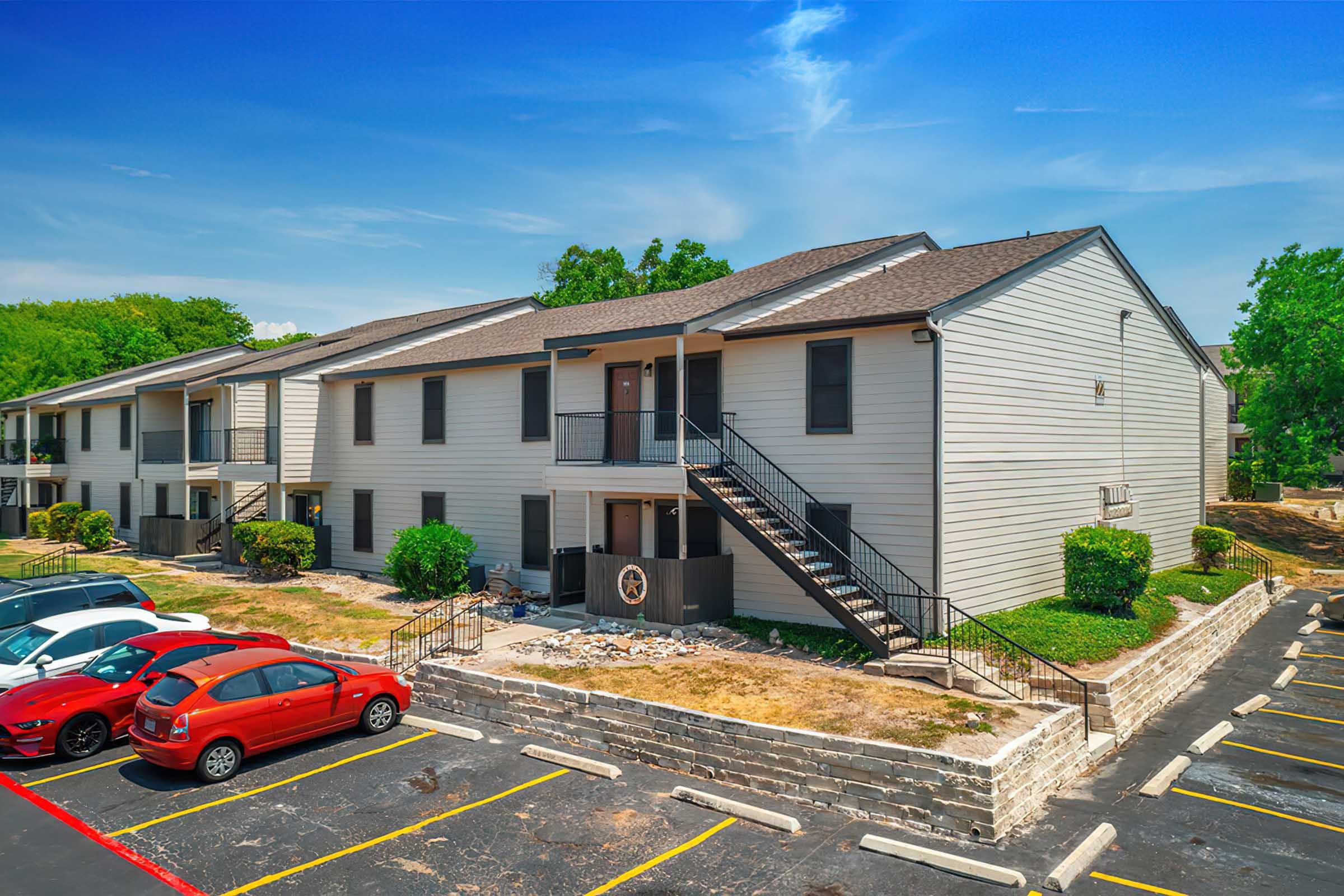 A view of a two-story apartment building featuring a staircase, greenery, and a parking lot with several cars. The blue sky and trees in the background enhance the suburban atmosphere.