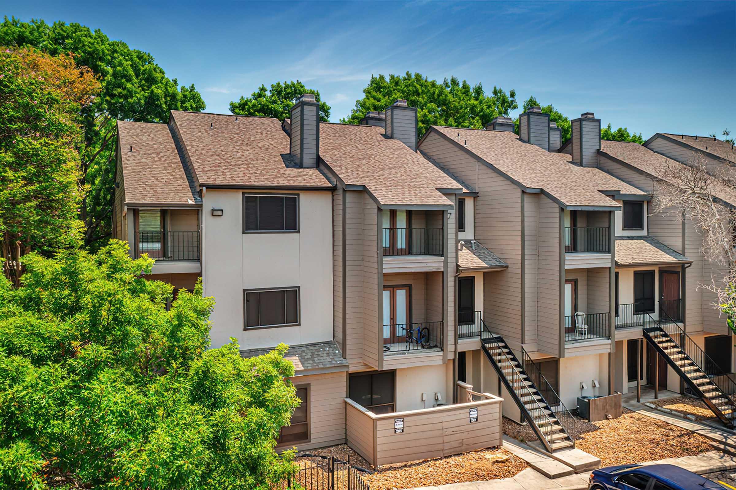 A view of a multi-unit residential building featuring multiple balconies and staircases. The exterior is a mix of light and dark siding, with landscaped greenery surrounding the property. Trees in the background suggest a well-maintained environment, and the sky is clear with minimal clouds.