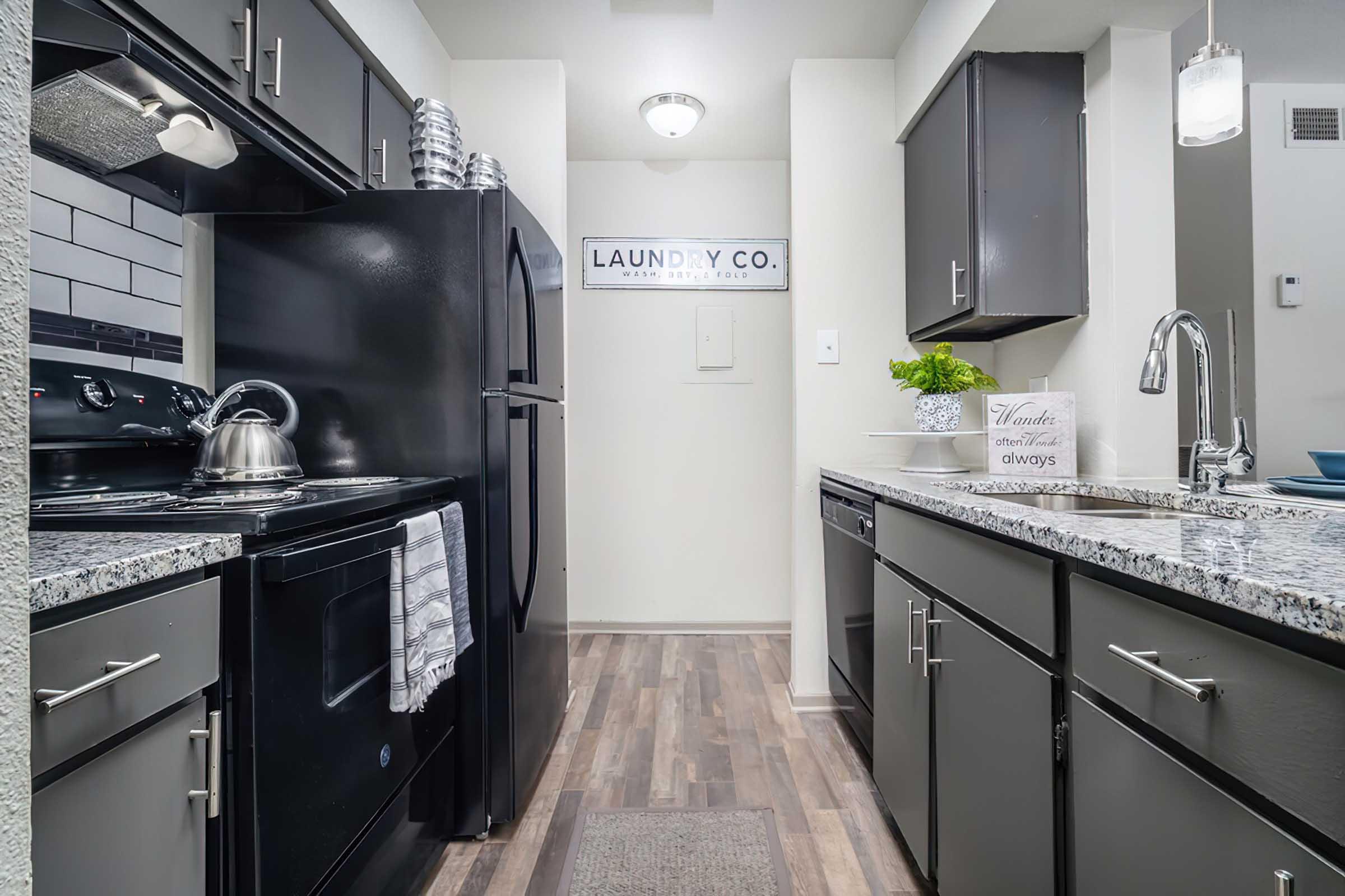 Modern kitchen featuring dark cabinetry, a granite countertop, and stainless steel appliances. A kettle is on the stove, and a laundry-themed sign hangs on the wall. The flooring is light wood, and the space is well-lit with a ceiling fixture, creating a welcoming atmosphere.