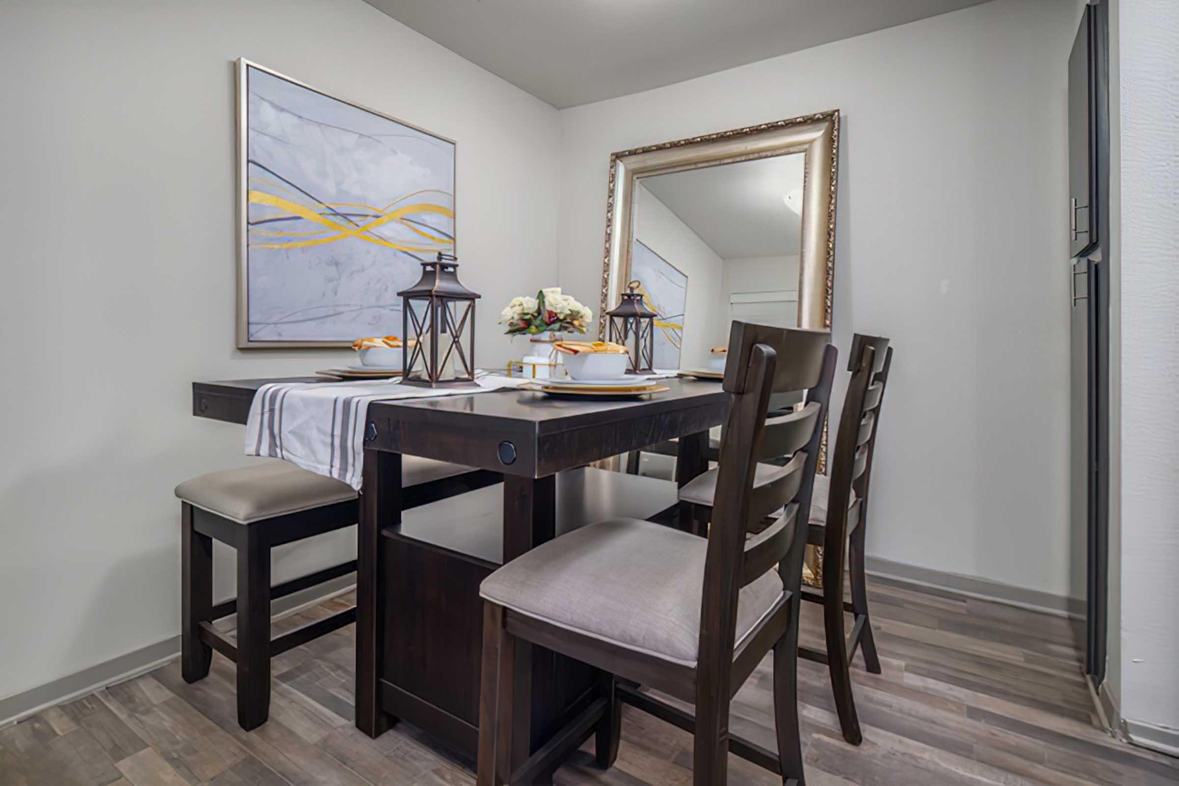 A modern dining area featuring a dark wooden table with high-backed chairs, set with plates and decorative items. A large mirror and abstract artwork hang on the wall, adding elegance to the space. The floor has a sleek, contemporary wood finish, creating a welcoming atmosphere.