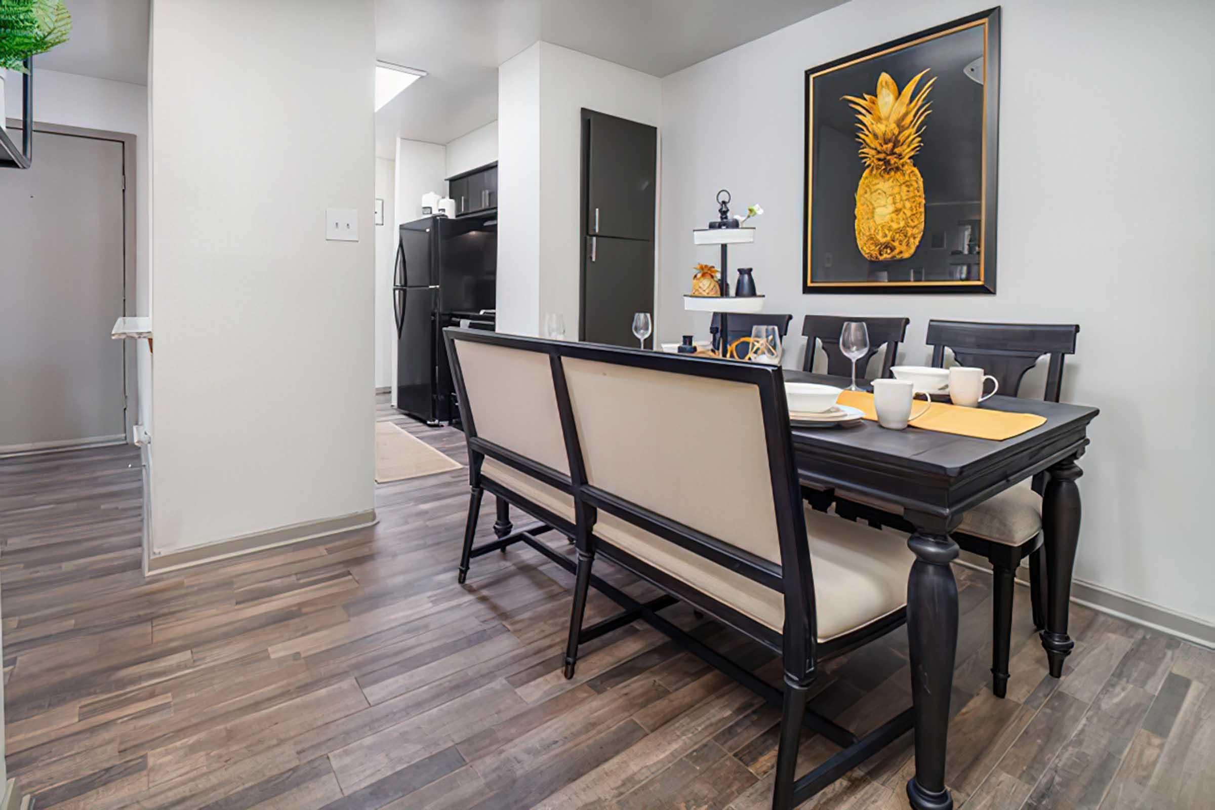 A modern dining area featuring a dark wooden table set for four with plates, glasses, and utensils. In the background, there is a sleek black refrigerator and a kitchen with white cabinetry. A framed artwork of a pineapple hangs on the wall, and the flooring is light-colored wood. Natural light enters the space, creating a welcoming ambiance.