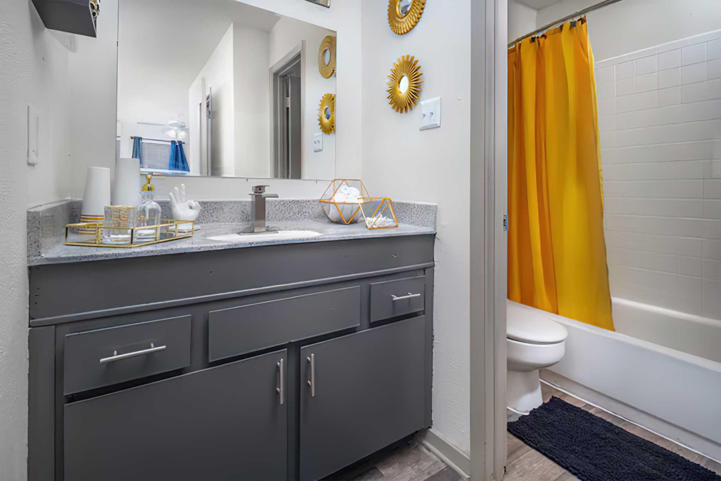 A modern bathroom featuring a gray vanity with a sink, decorative gold accents, a shower curtain in bright yellow, and a white bathtub. The space is well-lit, showcasing a clean and stylish design with a navy blue bath mat on the floor.