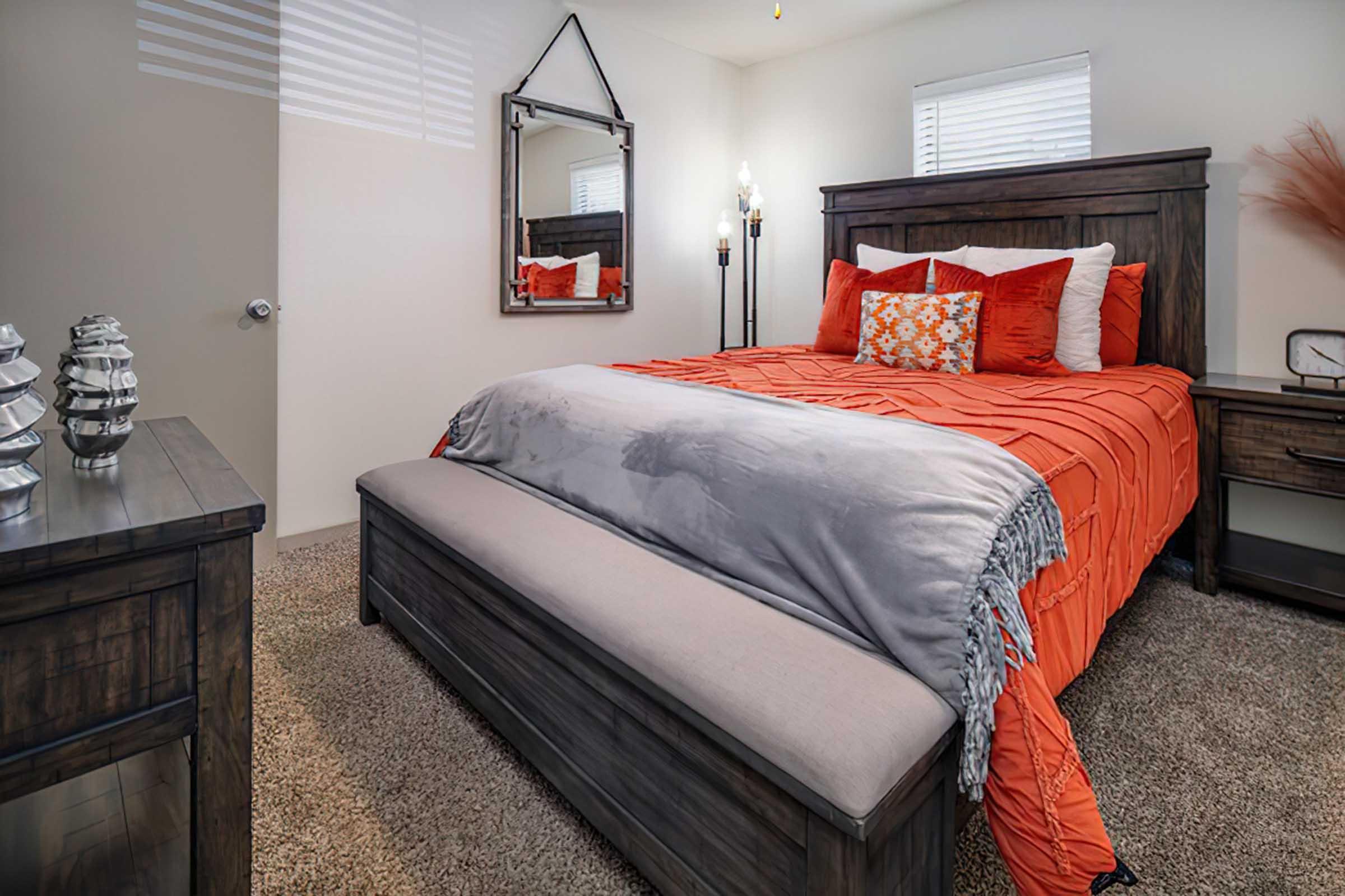 A cozy bedroom featuring a wooden bed with an orange and gray bedspread, flanked by two decorative pillows. There's a wooden nightstand with decorative items, a stylish mirror on the wall, and soft carpeting. Natural light filters through the window, creating a warm ambiance.