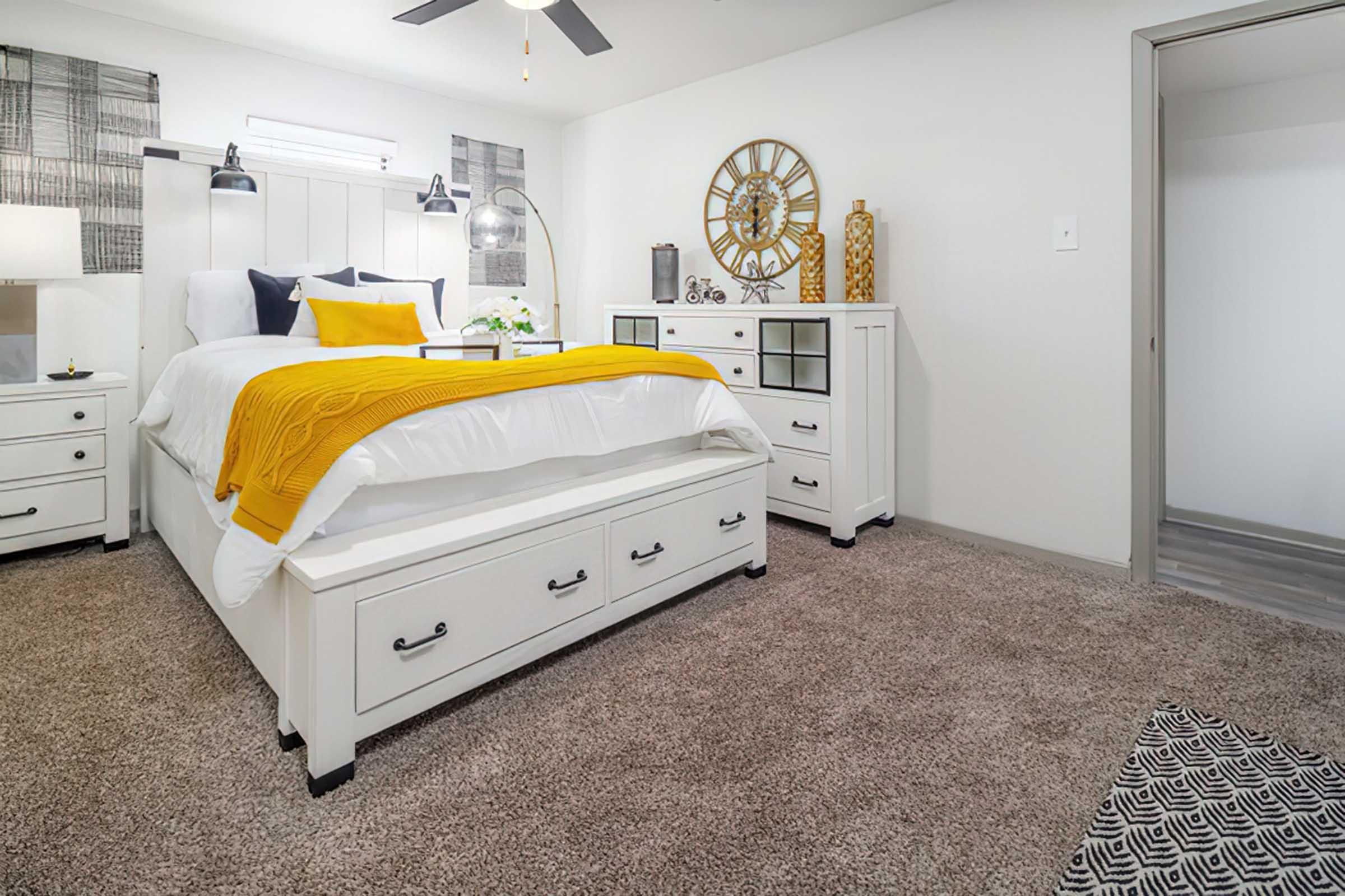 A cozy bedroom featuring a white bed with yellow and gray accents, bedside tables with lamps, a decorative wall clock, and a textured area rug. The walls are light-colored, and there's a ceiling fan for ventilation, creating a warm and inviting atmosphere.