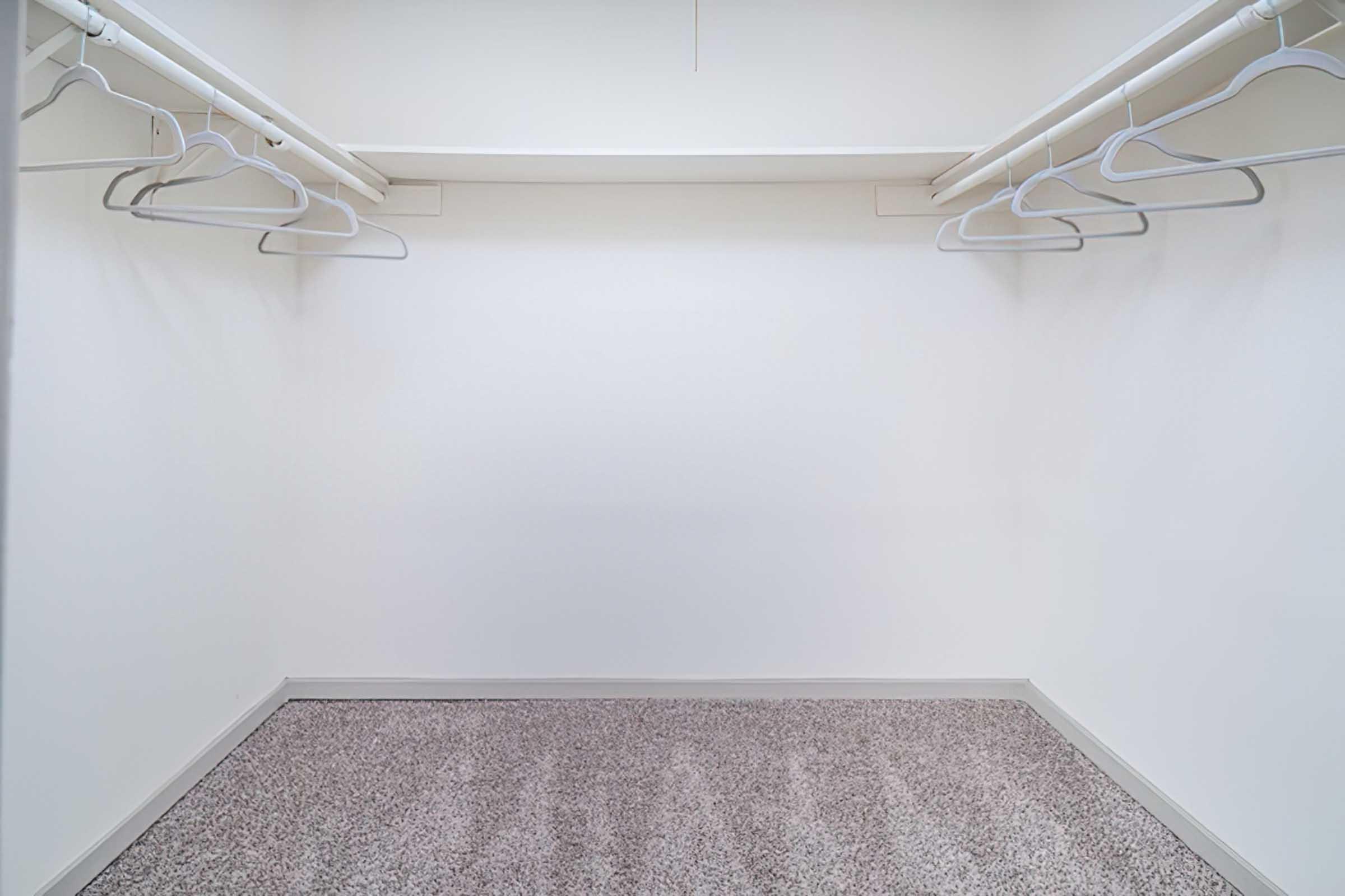 Empty closet with white walls and a carpeted floor. Several white hangers are hanging from a clothing rod, with ample space and no personal items visible. The overall atmosphere is clean and minimalistic.