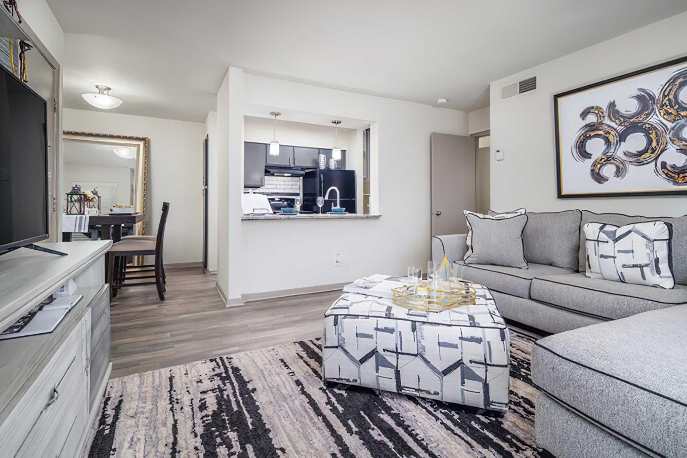 A modern living room featuring a gray sectional sofa with decorative pillows, a geometric coffee table, and a large area rug. In the background, a dining area with a wooden table and chairs is visible, along with a kitchen showcasing dark cabinets and stainless steel appliances. The space is well-lit and stylishly decorated.