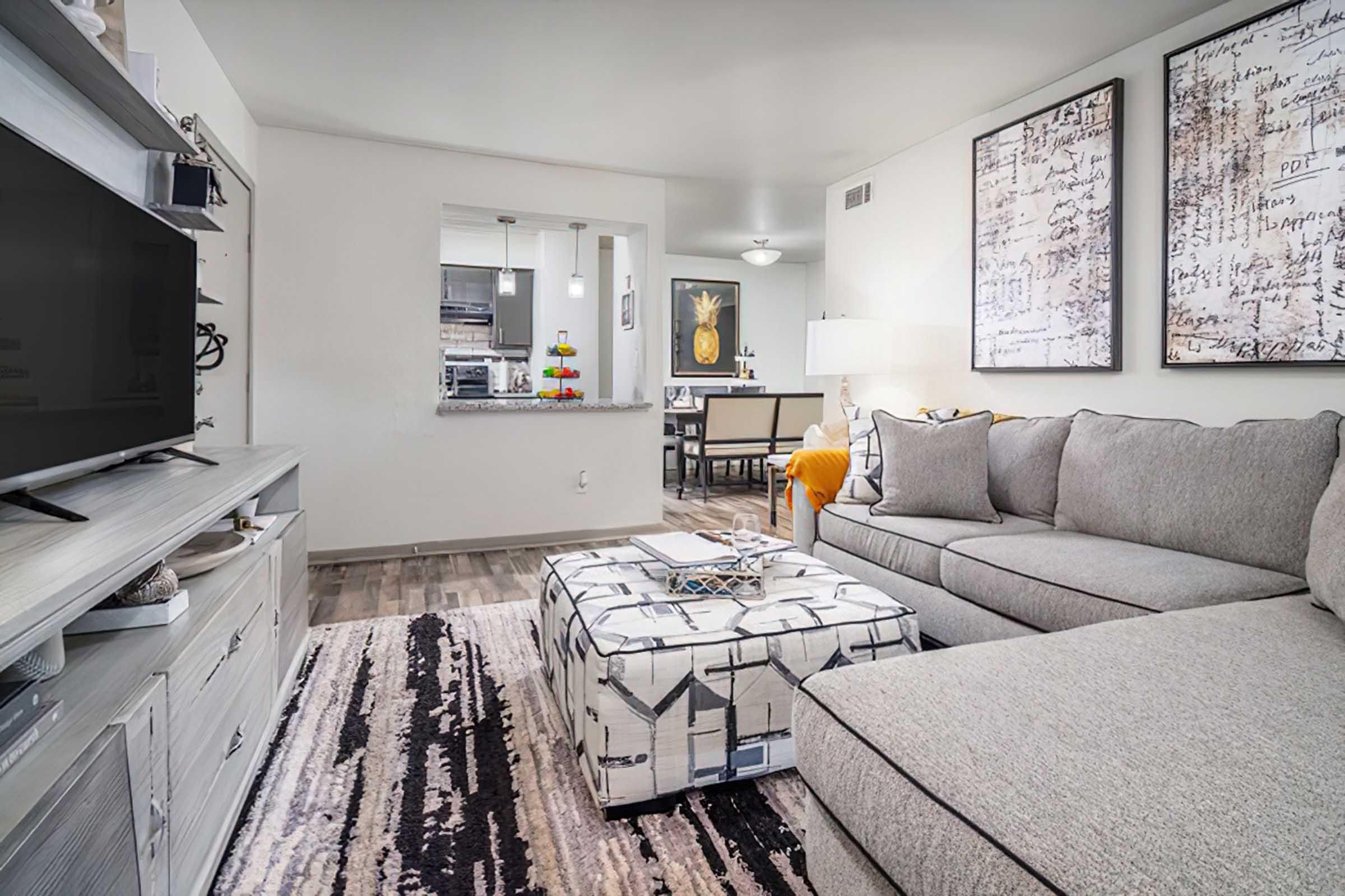 A modern living room featuring a gray sectional sofa, an ottoman with a geometric pattern, and a large TV stand. The walls are adorned with abstract art pieces. A dining area is visible in the background with a table and chairs. The space is well-lit, with a cozy and contemporary atmosphere.