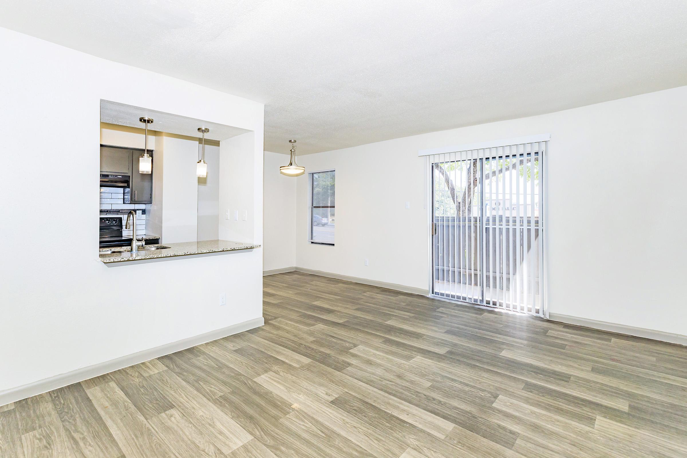A spacious, modern living area featuring laminate flooring, a large window with vertical blinds, and an open kitchen with a granite countertop and pendant lighting. The walls are painted in a light color, creating a bright and inviting atmosphere.
