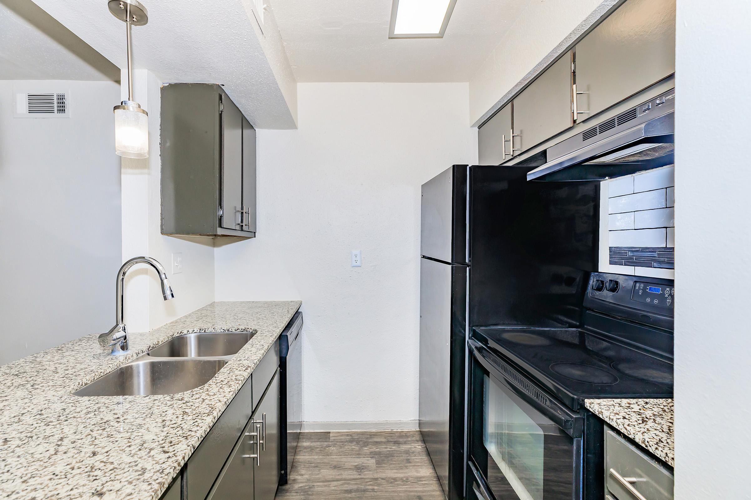 Modern kitchen featuring dark cabinetry, granite countertops, and stainless steel appliances. Includes a double sink, built-in oven, and black refrigerator. The space is well-lit with recessed lighting and a pendant light hanging above the kitchen island. The flooring is a light wood design.