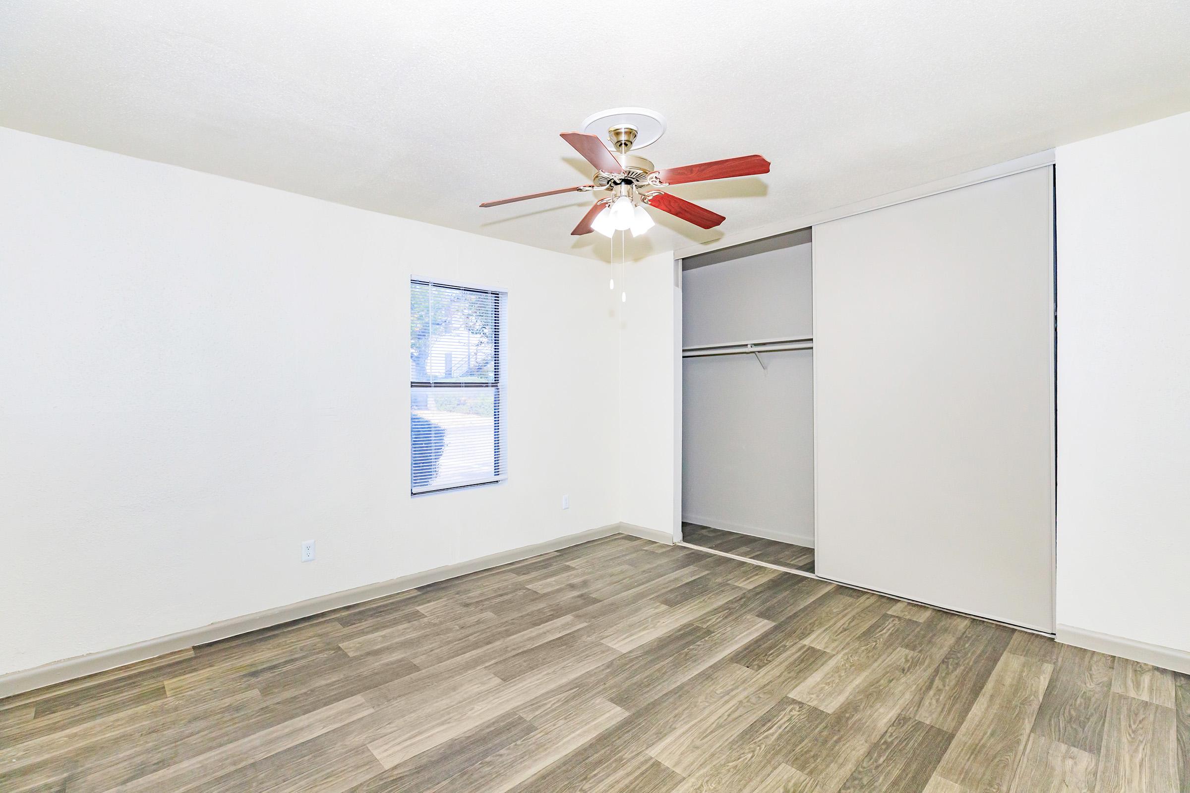 Bright, empty room with light-colored walls and a ceiling fan with red blades. Features a window with blinds and a sliding closet door. The floor is covered in wood-like tiles, creating a modern and clean appearance.