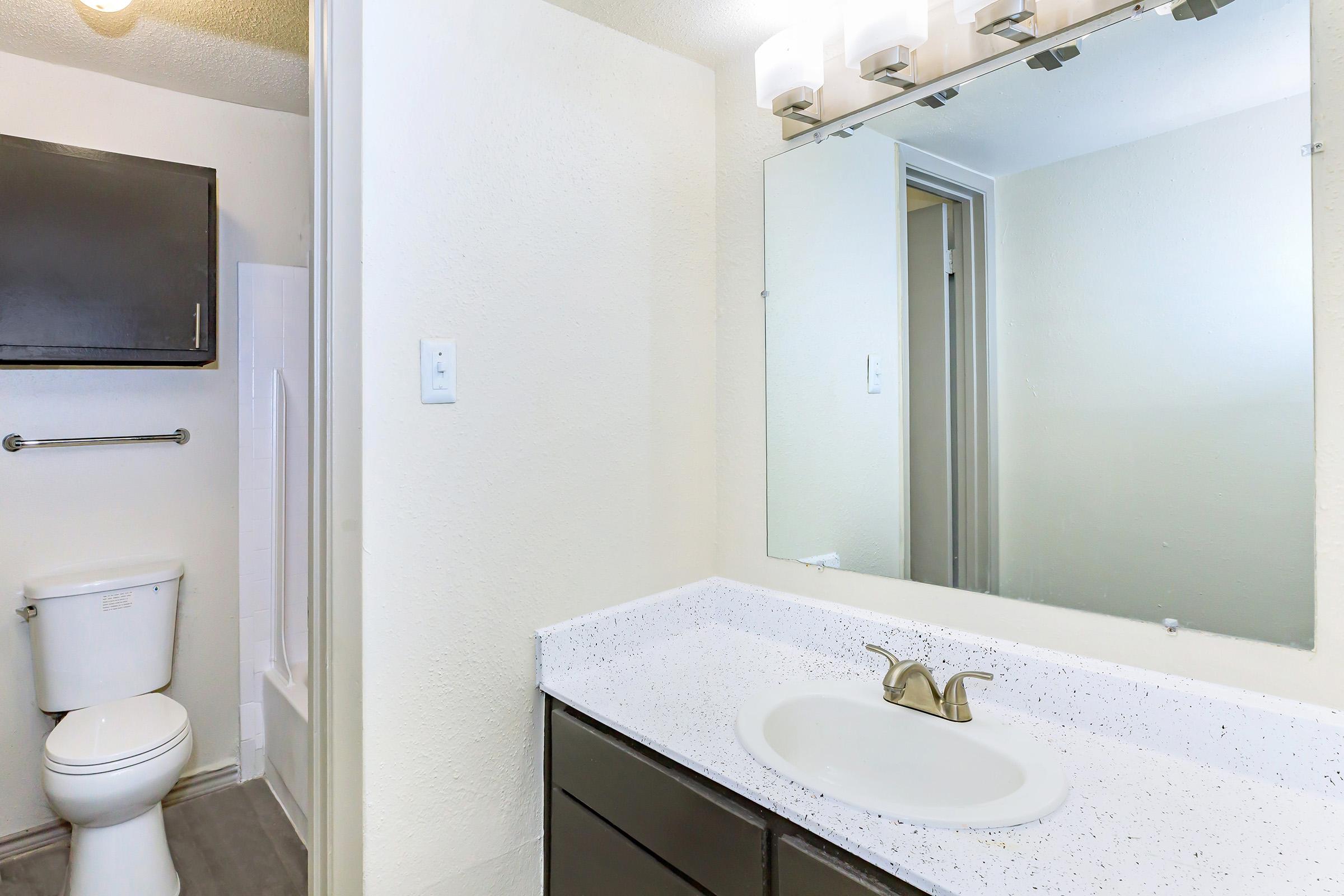 A modern bathroom featuring a white countertop with a sink, a large wall mirror, light fixtures above, a dark cabinet, a toilet, and a shower area with white tiles. The walls are painted in light colors, creating a bright and clean atmosphere.