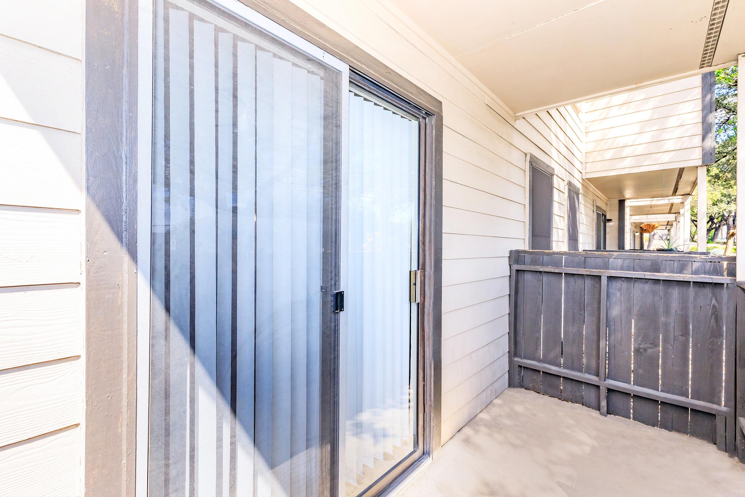 A glass sliding door with vertical blinds leading to a small, enclosed patio area. The patio is bordered by a wooden fence, and in the background, there are more apartment units visible, partially obscured by trees. The scene is bright, suggesting a sunny day.
