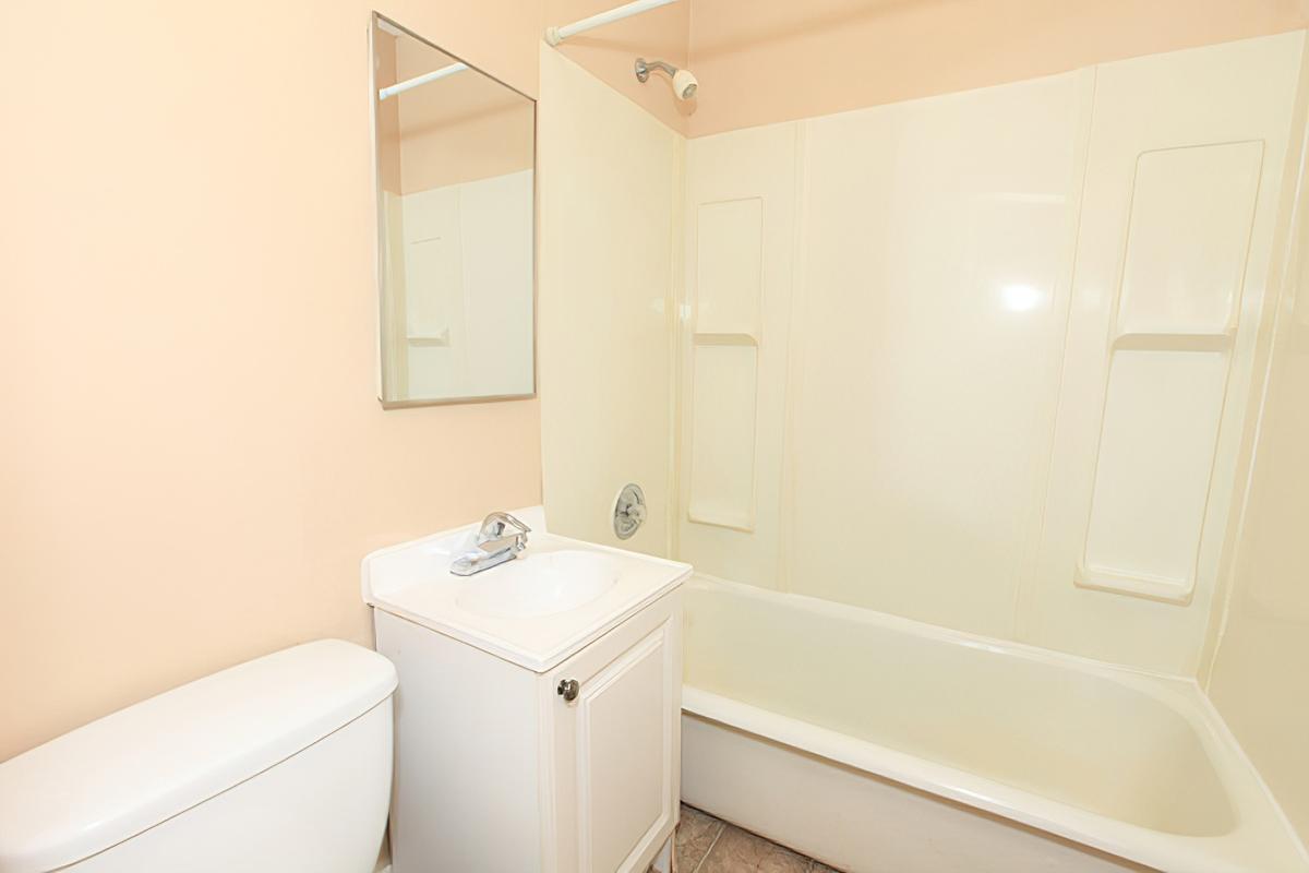 A clean, modern bathroom featuring a white bathtub with a shower, a small vanity with a sink, and a wall-mounted mirror. The walls are painted a light peach color, and the floor is tiled. Overall, the space appears bright and well-maintained.