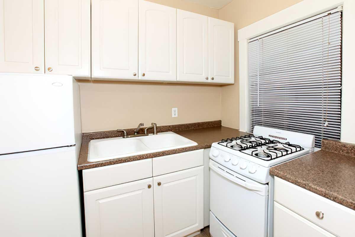 A small kitchen featuring white cabinetry, a double basin sink, a white refrigerator, and a gas stove with a visible oven below. The countertop is a dark color, and there is a window with horizontal blinds providing natural light. The walls are painted a light beige.