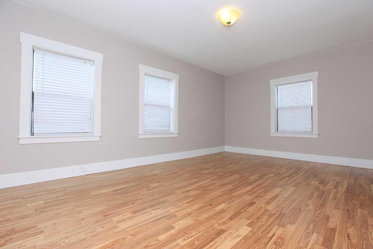 Empty room with light-colored walls and three windows with white blinds. The floor is made of light wood, creating a spacious and bright atmosphere. A ceiling light fixture is visible, providing illumination. The room is free of furniture, highlighting its size and potential for various uses.