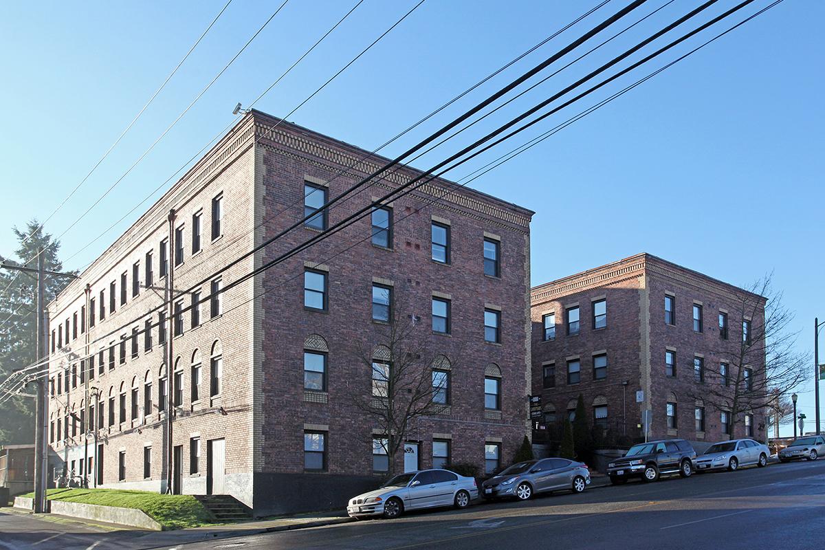 Two brick apartment buildings stand side by side against a clear blue sky, with multiple windows on each level. Power lines stretch across the image above the buildings. Several cars are parked along the street in front. The scene conveys an urban residential environment.