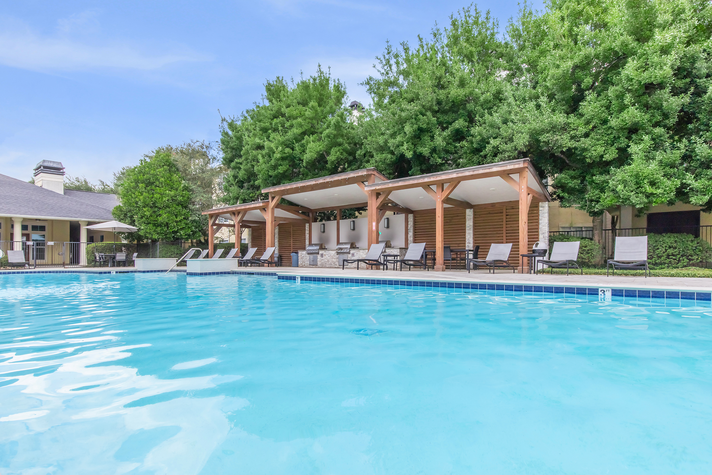 A serene swimming pool surrounded by lounge chairs and shaded cabanas. Lush green trees line the background, providing a natural setting. The water is clear and reflects the blue sky above, creating a relaxing atmosphere ideal for leisure and enjoyment.