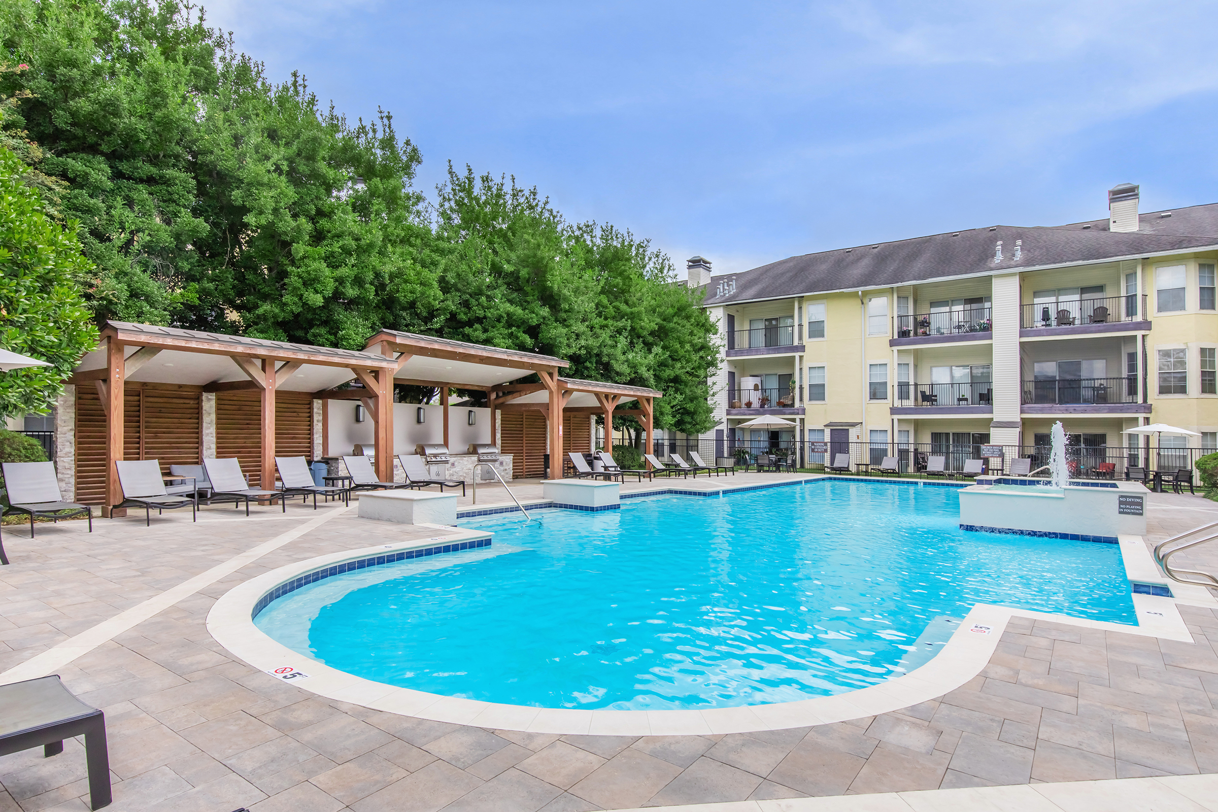A serene outdoor swimming pool surrounded by lounge chairs and shaded gazebo areas, set against a backdrop of green trees and a multi-story residential building. The water is calm and inviting, perfect for relaxation on a sunny day.