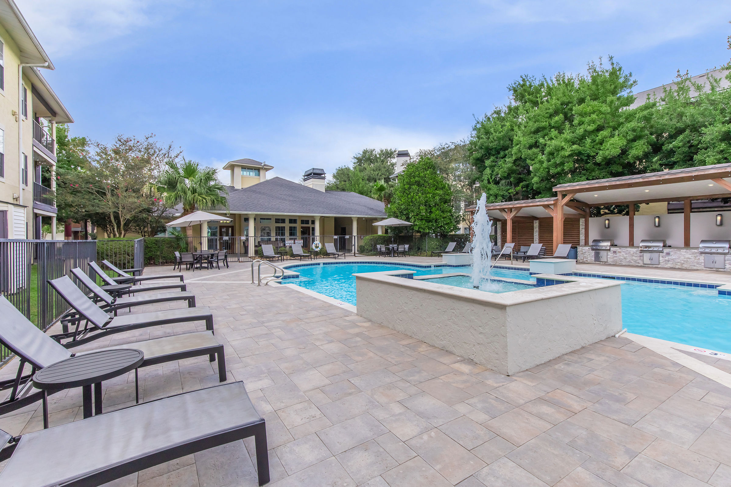 Swimming pool area featuring a fountain, lounge chairs, and shaded cabanas. The modern building in the background is surrounded by lush greenery and trees. The sky is clear with a light blue hue, creating a relaxing outdoor atmosphere.