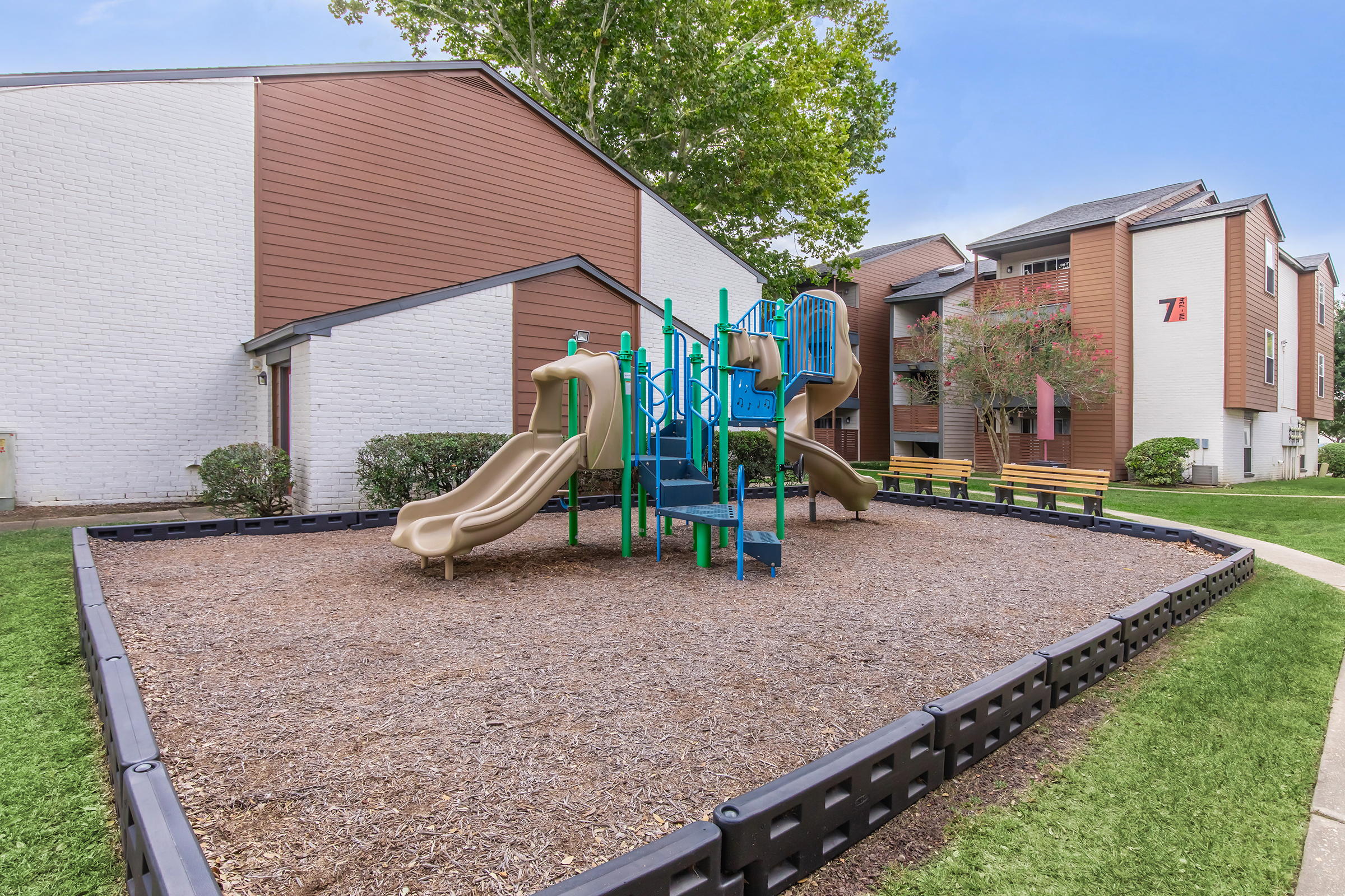 Playground area with two slides and a climbing structure surrounded by soft mulch. The playground is located next to a multi-unit residential building with greenery and benches nearby, under a clear blue sky.