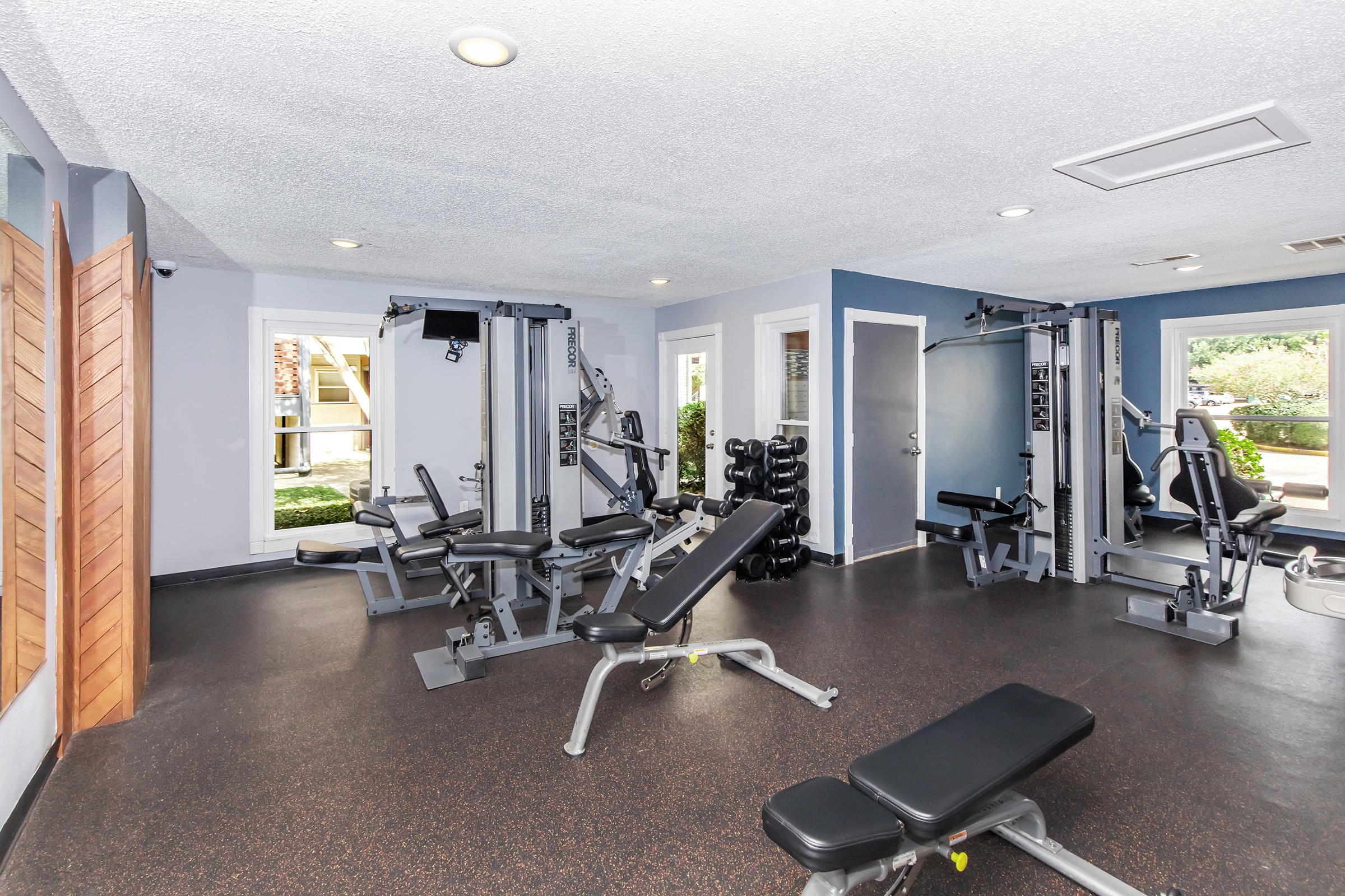 A modern fitness room featuring various gym equipment, including weight machines, benches, and free weights. Large windows let in natural light, creating a bright atmosphere. The flooring is dark and padded for comfort, and the walls are painted in a calming blue color.
