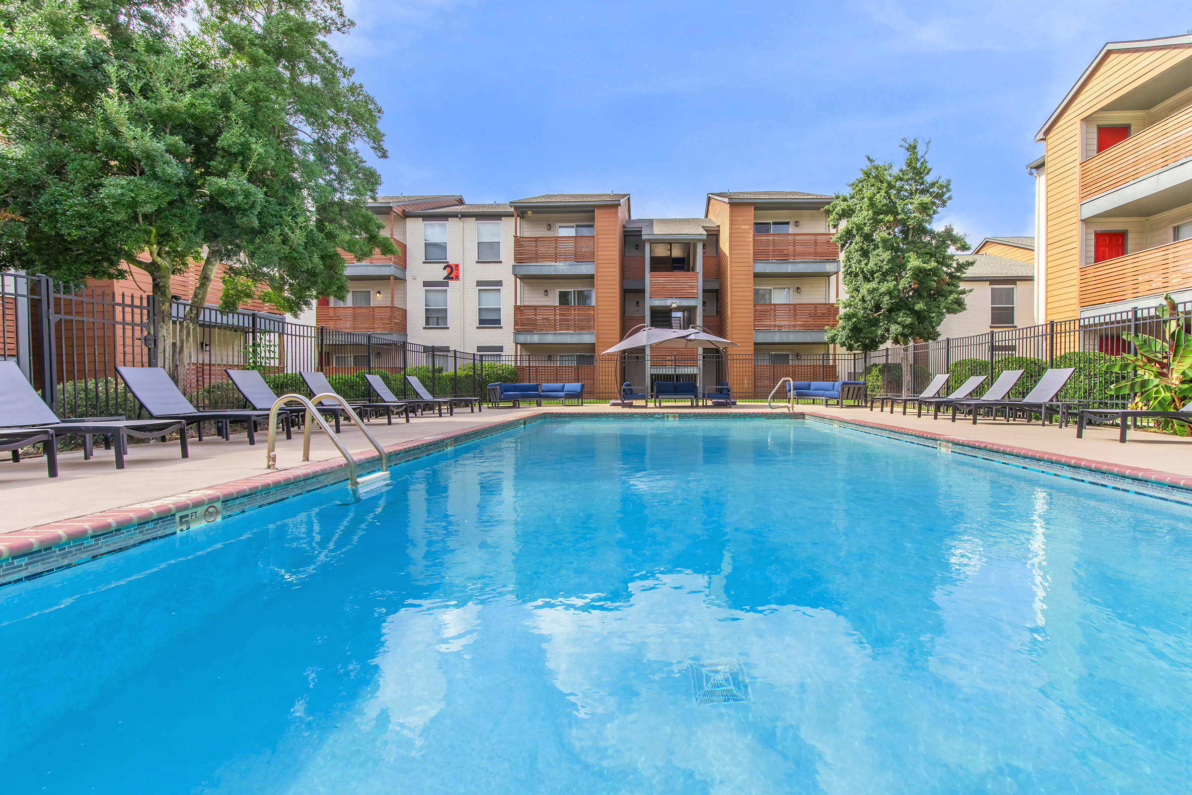 A clear blue swimming pool surrounded by lounge chairs, with trees and a multi-story apartment building in the background. The sky is bright and sunny, creating a relaxing atmosphere. The area is well-maintained, perfect for leisure and social gatherings.