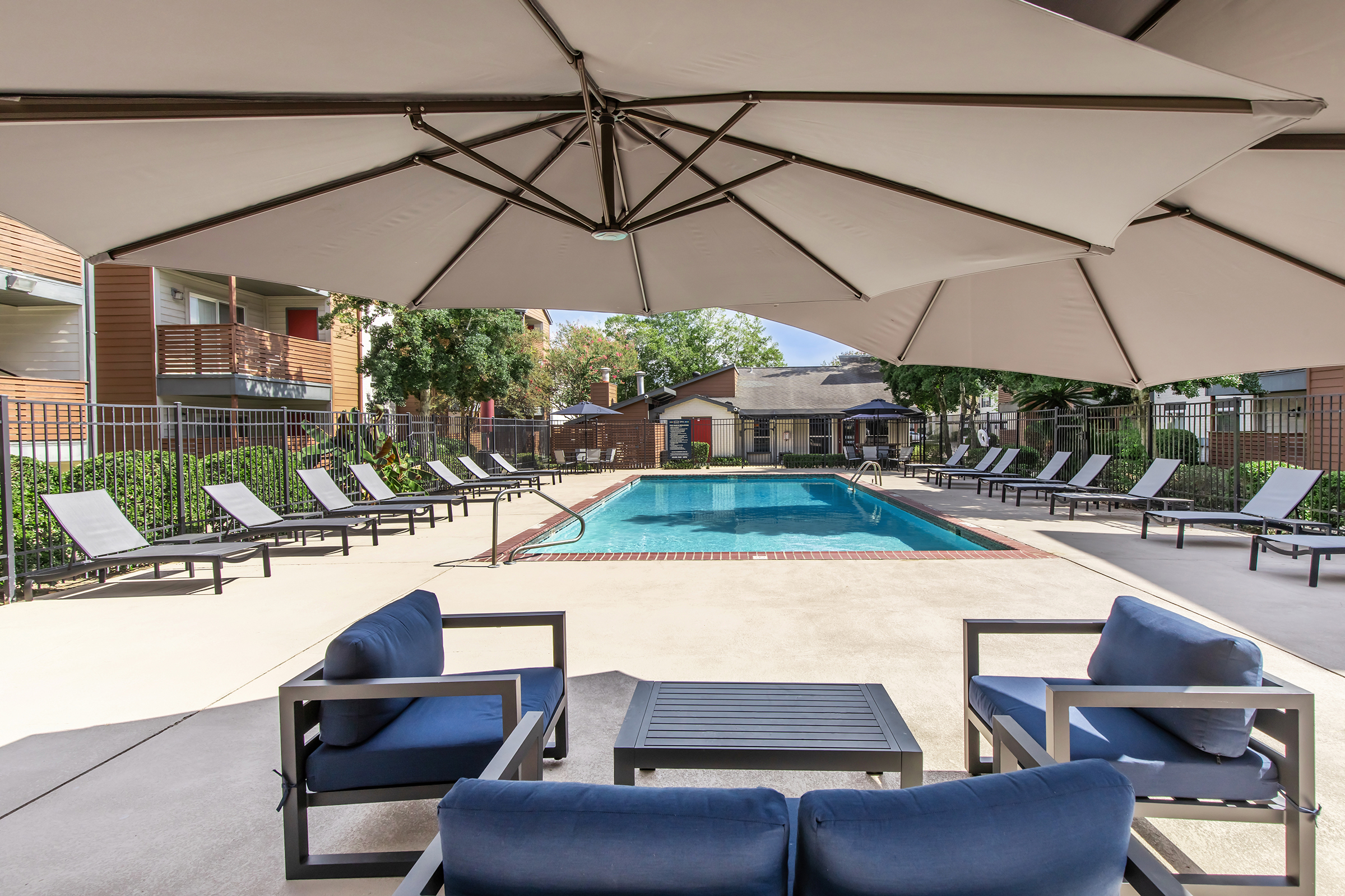 A well-maintained outdoor pool area surrounded by lounge chairs and shaded by large umbrellas. The space includes a few small tables and is enclosed by a fence, with landscaped greenery in the background. Ideal for relaxation and recreation.