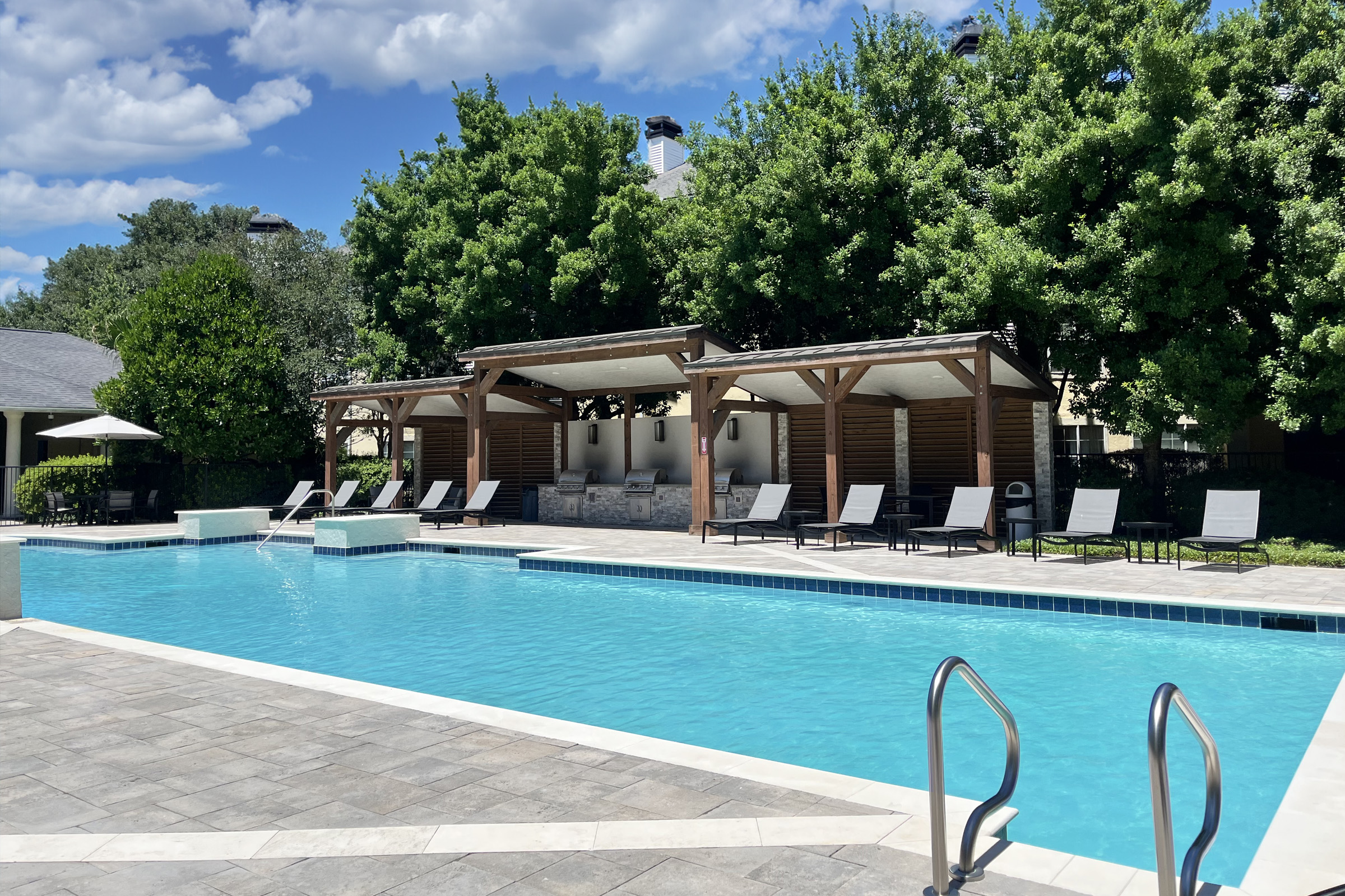 A tranquil outdoor swimming pool surrounded by green trees and lounging chairs. In the background, a wooden pergola provides shaded seating. The clear blue water reflects the bright sky with fluffy white clouds, creating a serene and inviting atmosphere.