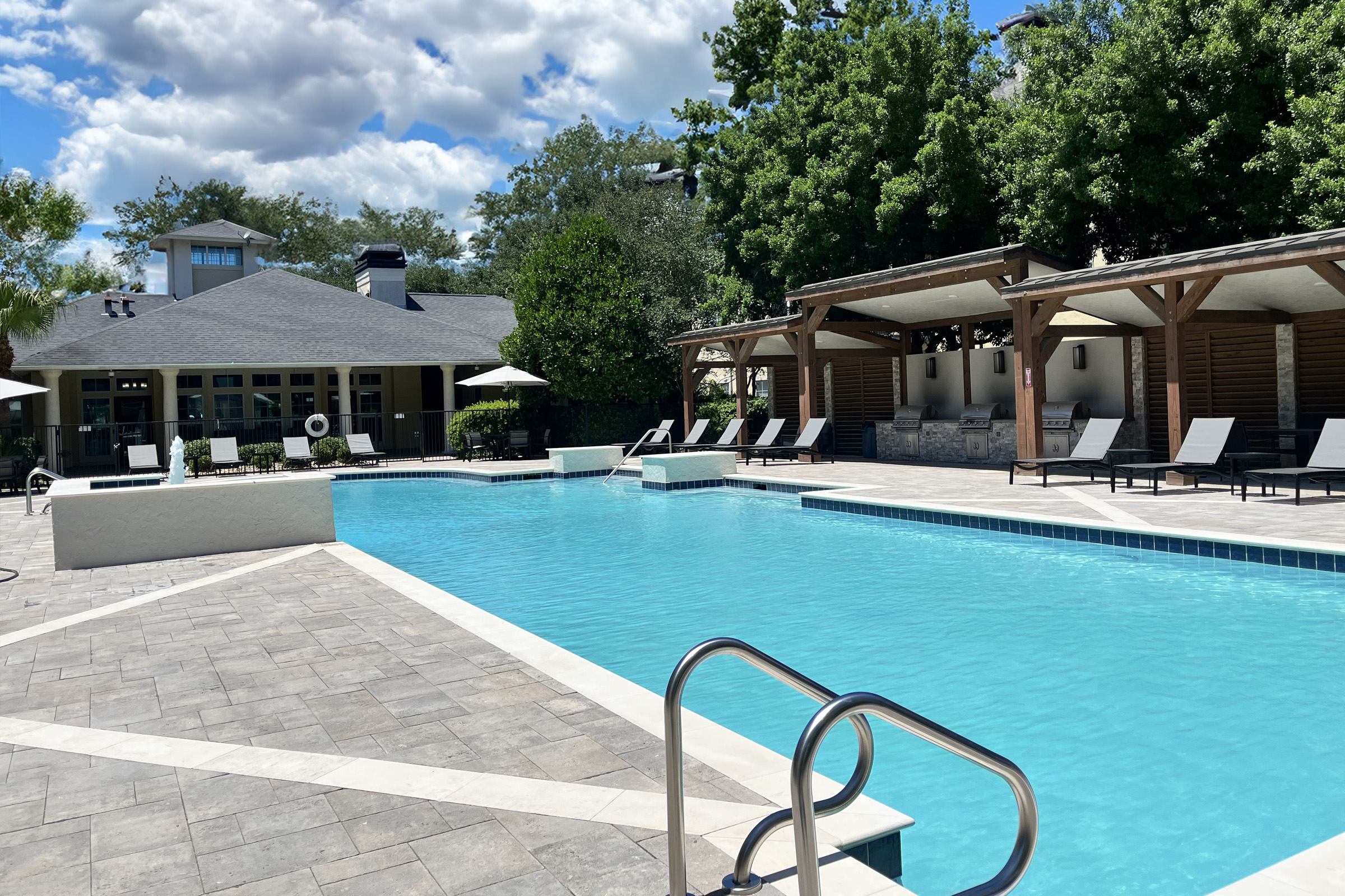 A shimmering outdoor swimming pool with a decorative edge, surrounded by lounge chairs and shaded cabanas. In the background, there is a modern building with large windows and a landscaped area with trees under a partly cloudy sky.