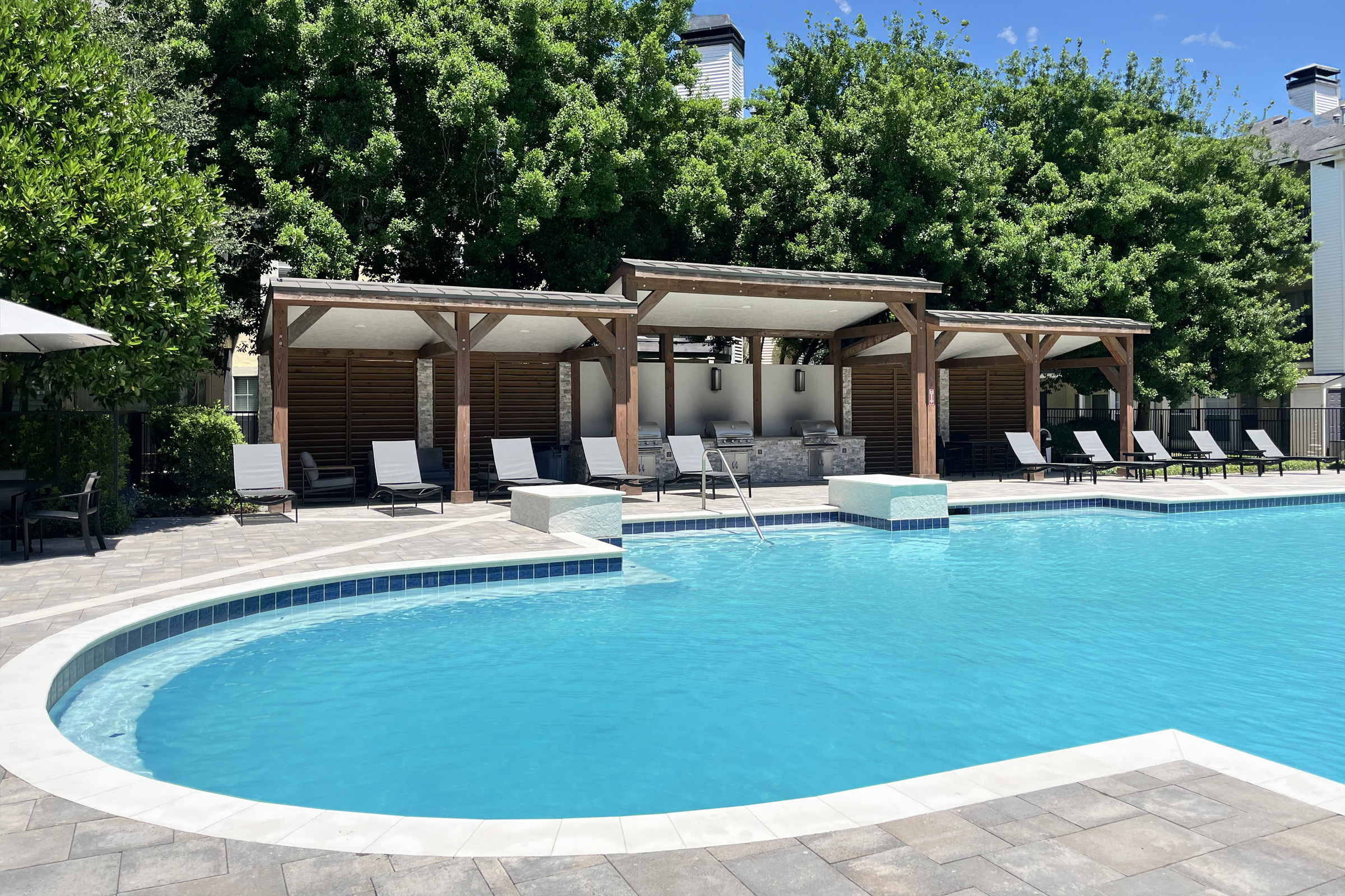 A bright and inviting outdoor pool area featuring clear blue water, surrounded by lounge chairs and shaded cabanas. Lush green trees provide a natural backdrop, and a sunny sky enhances the inviting atmosphere. The pool deck is paved with light-colored stones, creating a relaxing setting for leisure and enjoyment.