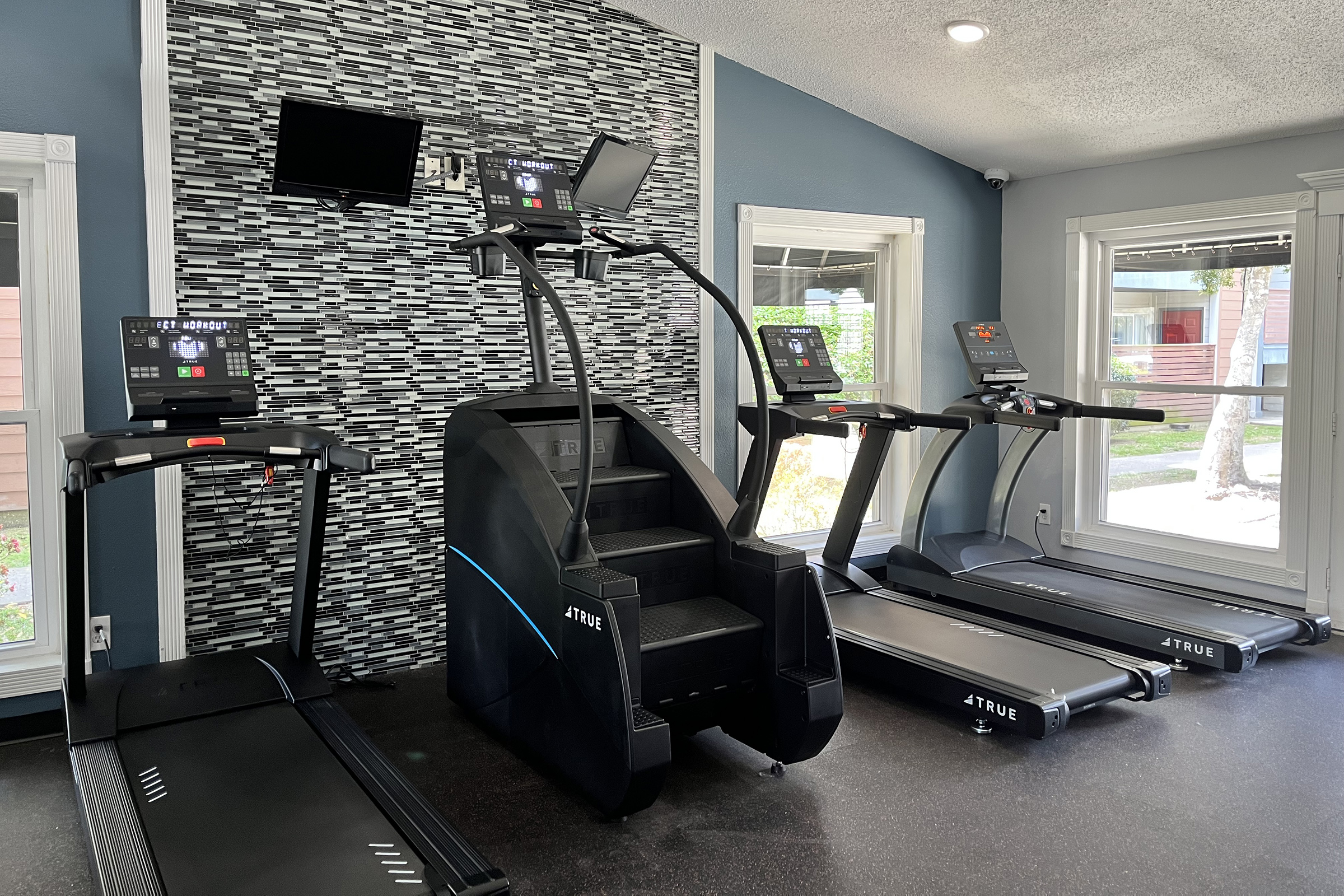 A modern gym interior featuring several fitness machines, including treadmills and a stair climber. The machines have digital screens displaying information. The walls are painted blue, and there's a decorative wall in the background. A window allows natural light to enter the space.