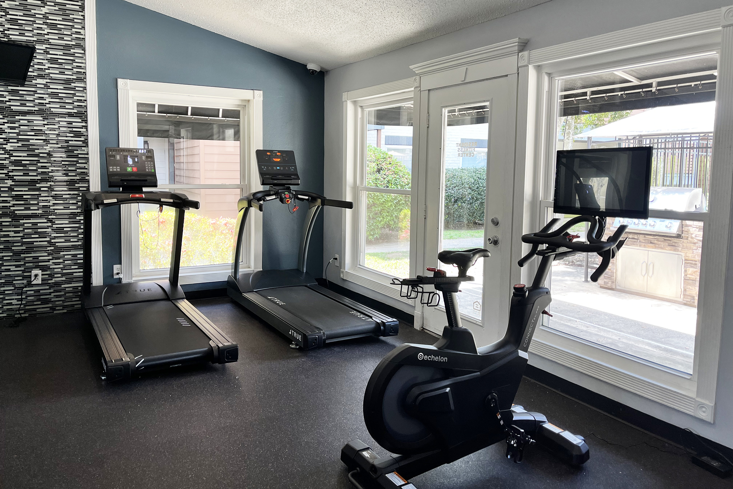 A fitness room featuring two treadmills and a stationary bike, all equipped with screens. Large windows allow natural light to enter, creating a bright atmosphere. The floor is dark, contrasting with the lighter walls and modern decor. The space appears well-maintained and inviting for workouts.