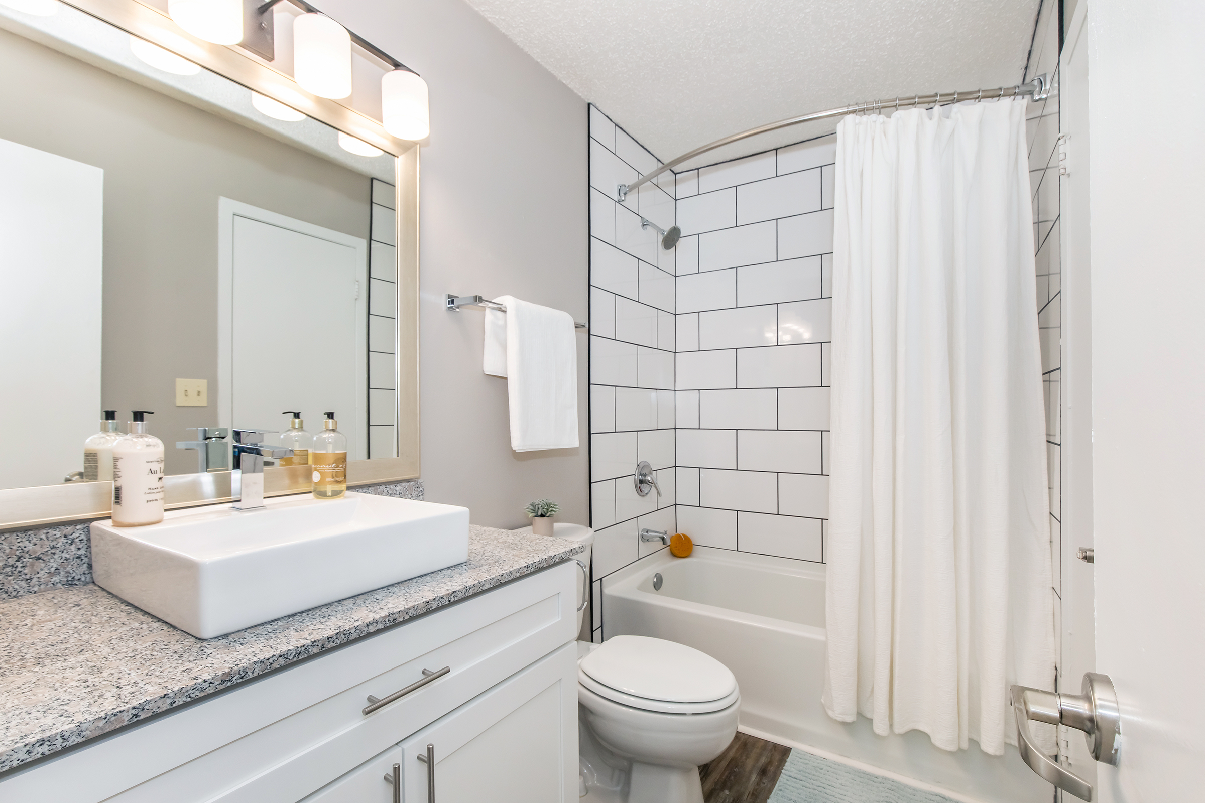 A modern bathroom featuring a white bathtub with a shower curtain, a rectangular sink on a granite countertop, and stylish lighting. The walls are tiled with white square tiles, and the space includes toiletries on the counter, a towel rack, and a small plant for decor.