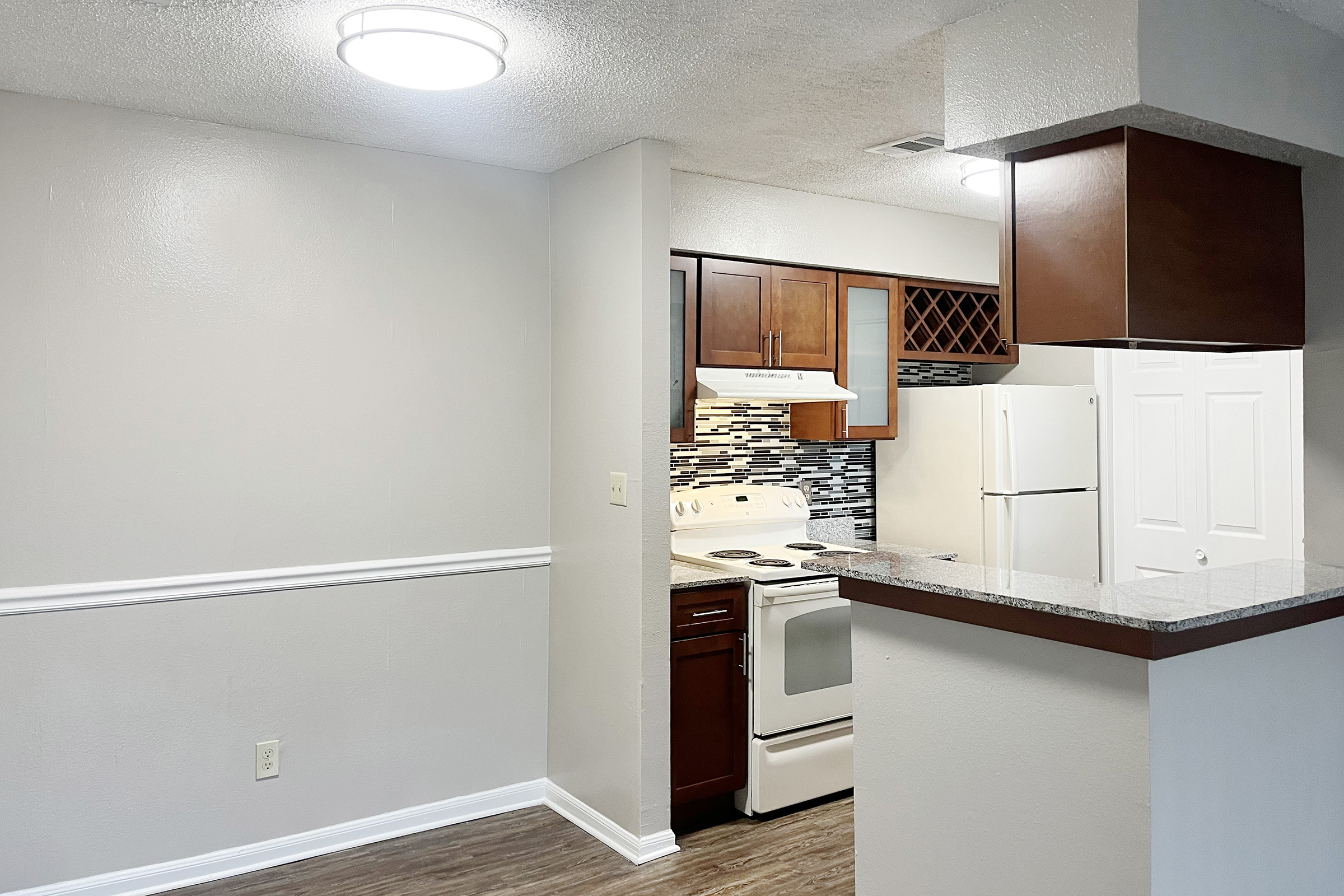 A modern kitchen featuring dark wood cabinets and a white stove, with a tile backsplash. There’s a refrigerator to the right. The adjacent living area has light gray walls, a textured ceiling, and a decorative white trim. Bright overhead lighting enhances the spacious feel.