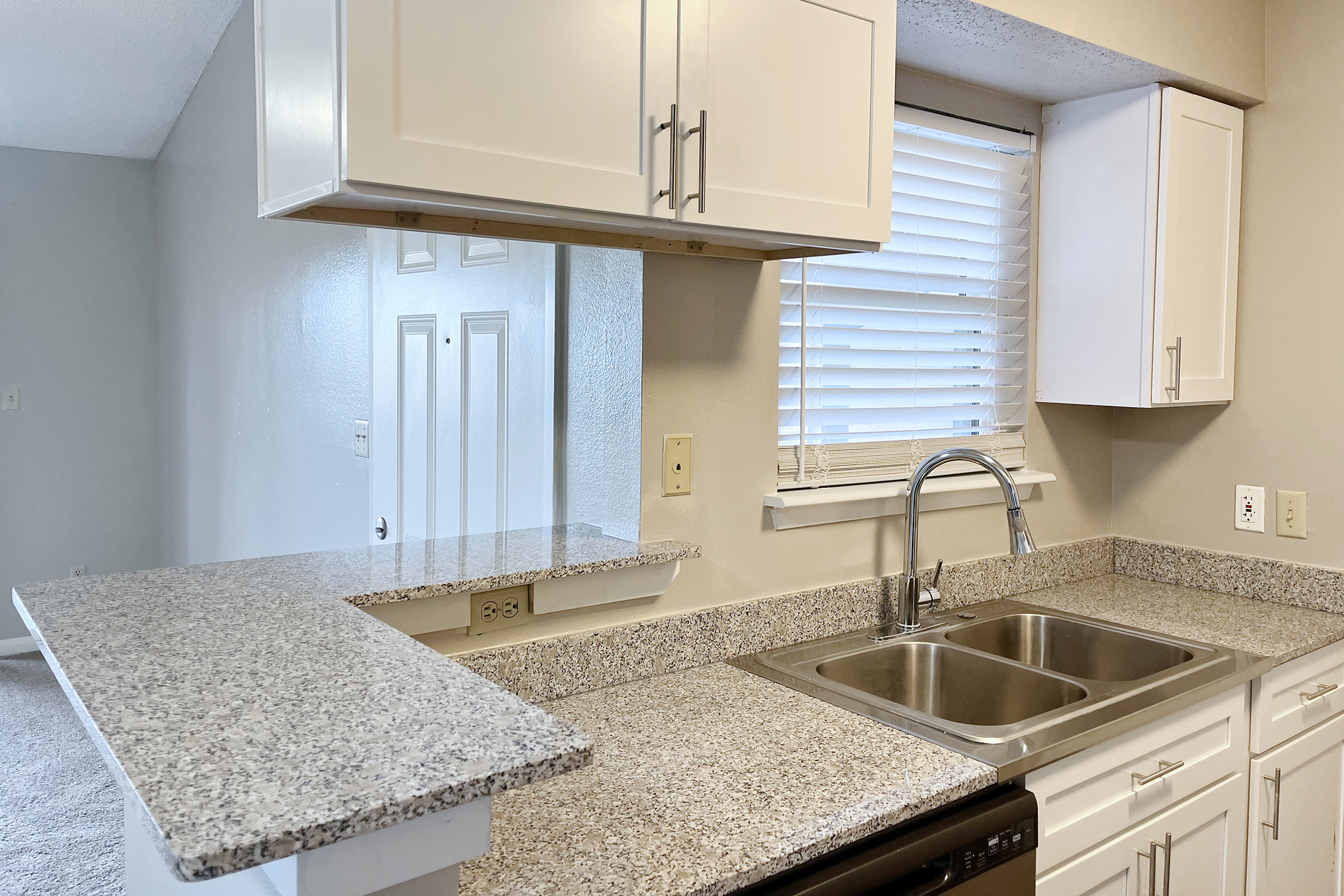 A modern kitchen featuring white cabinets, a granite countertop with a double sink, and a small breakfast bar. There are windows allowing natural light, and the space is connected to a living area with beige walls and carpet. A door is visible in the background.