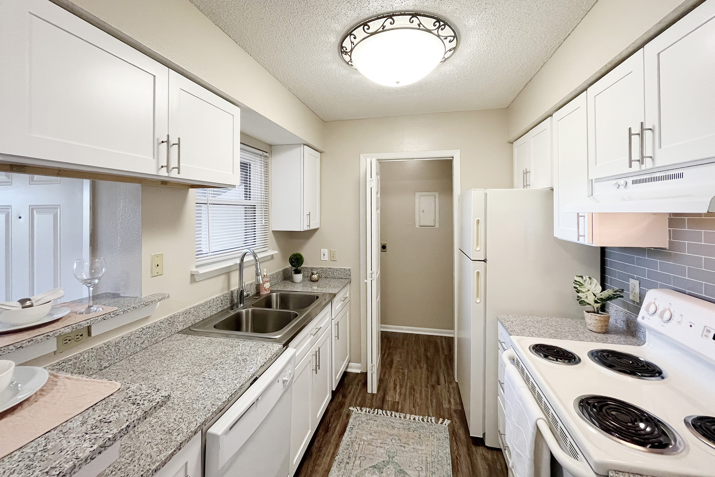 A modern kitchen featuring white cabinetry, a double sink, and granite countertops. The room is well-lit with a ceiling light and includes essential appliances like a refrigerator and stove. A small, decorative rug adds warmth, while a plant brings a touch of greenery, enhancing the inviting atmosphere.