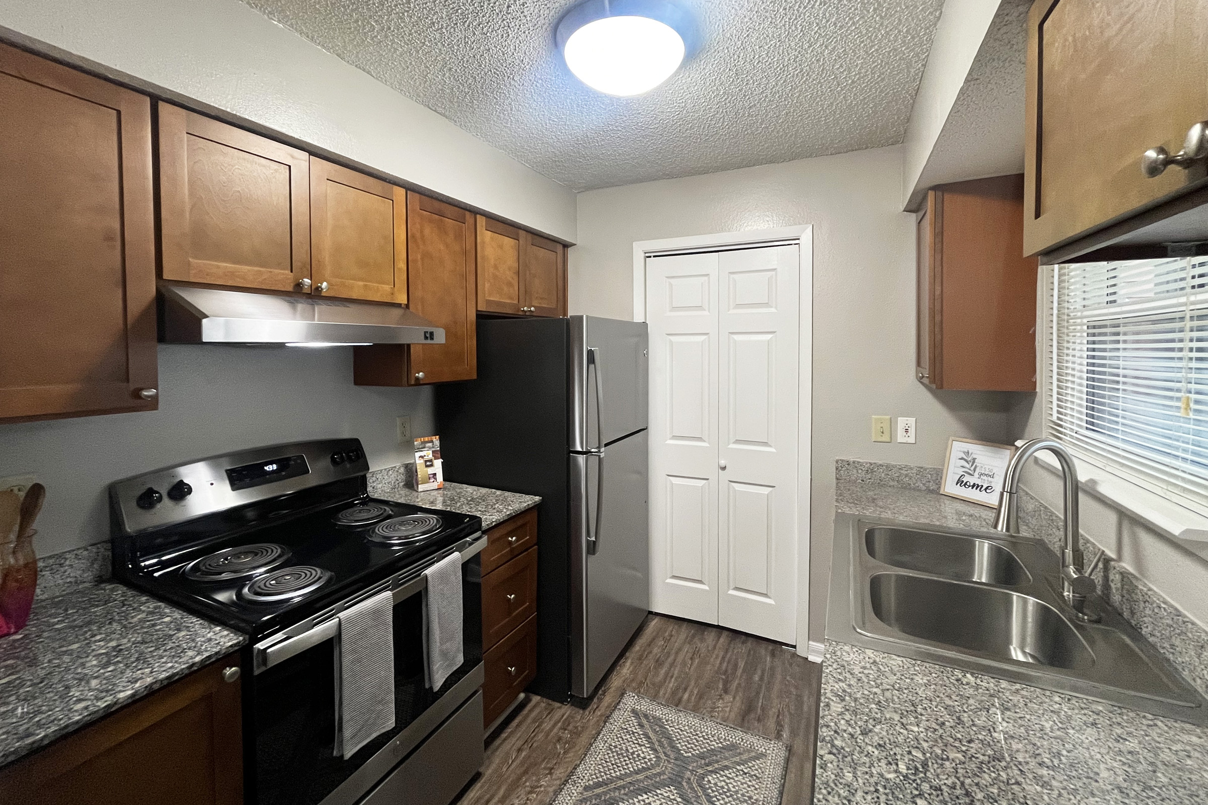 A modern kitchen featuring wooden cabinetry, a stainless steel refrigerator, and an electric stove with an oven. The countertops are granite, and there are two sinks next to the window. A door leads to a closet, and the room is well-lit with a ceiling fixture.