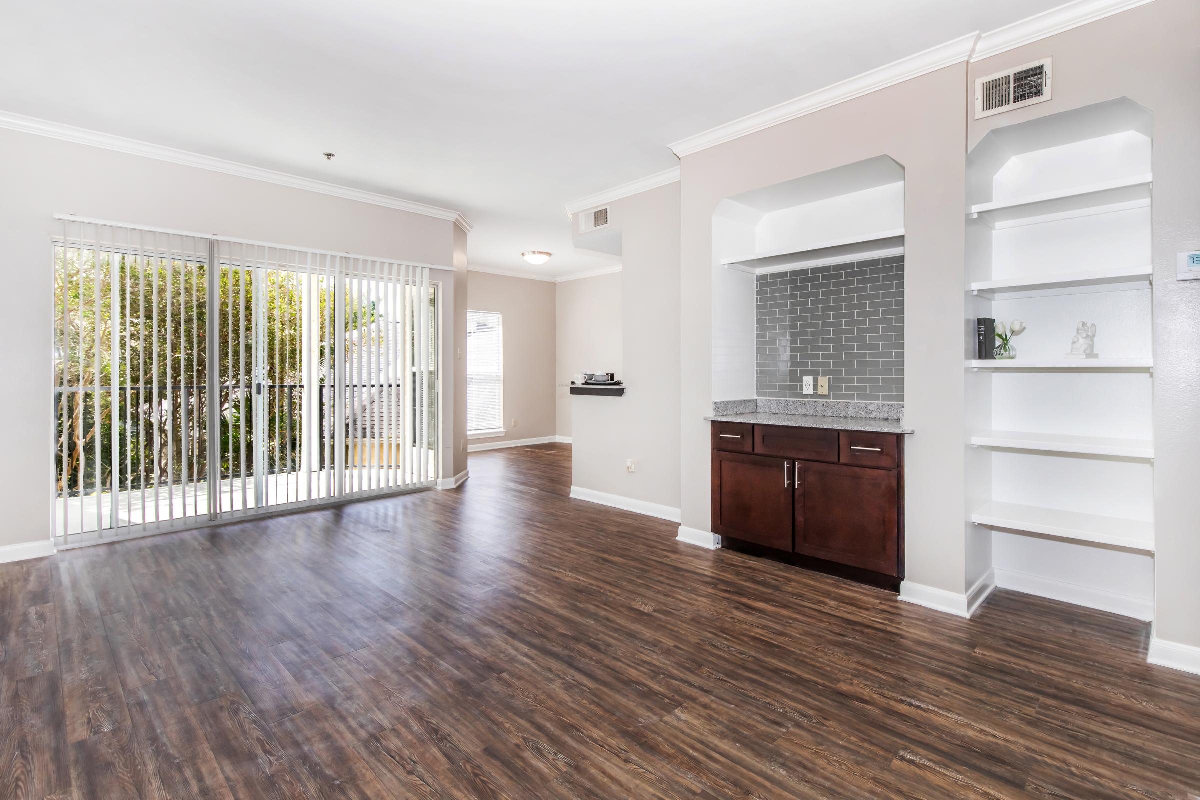 Bright and spacious living room with light-colored walls and wooden flooring. There are glass sliding doors leading to an outdoor area, allowing natural light to enter. A stylish kitchen nook features dark cabinets and a granite countertop, while a shelf unit is integrated into the design, enhancing the modern aesthetic.