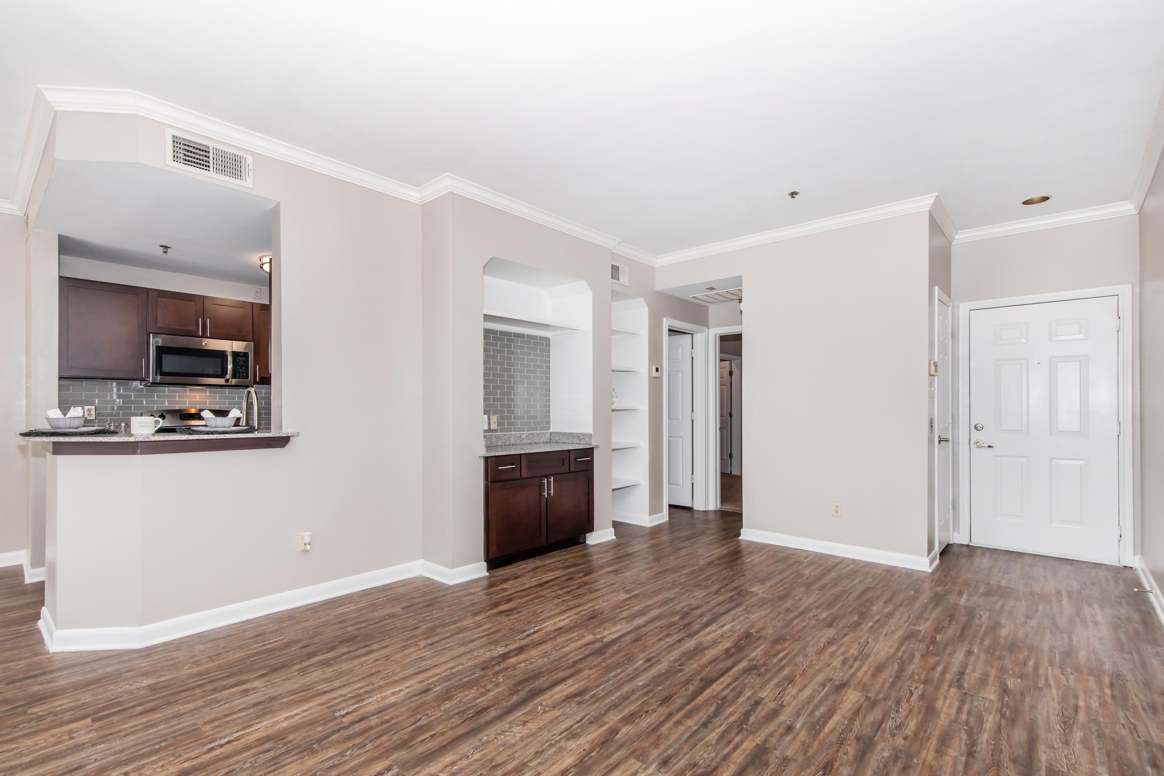 Spacious living area with light-colored walls and modern wood flooring. To the left, there’s a bar counter leading to a kitchen with dark cabinetry. A hallway leads to additional rooms, and a white door is visible on the right. Natural light brightens the space, creating an inviting atmosphere.