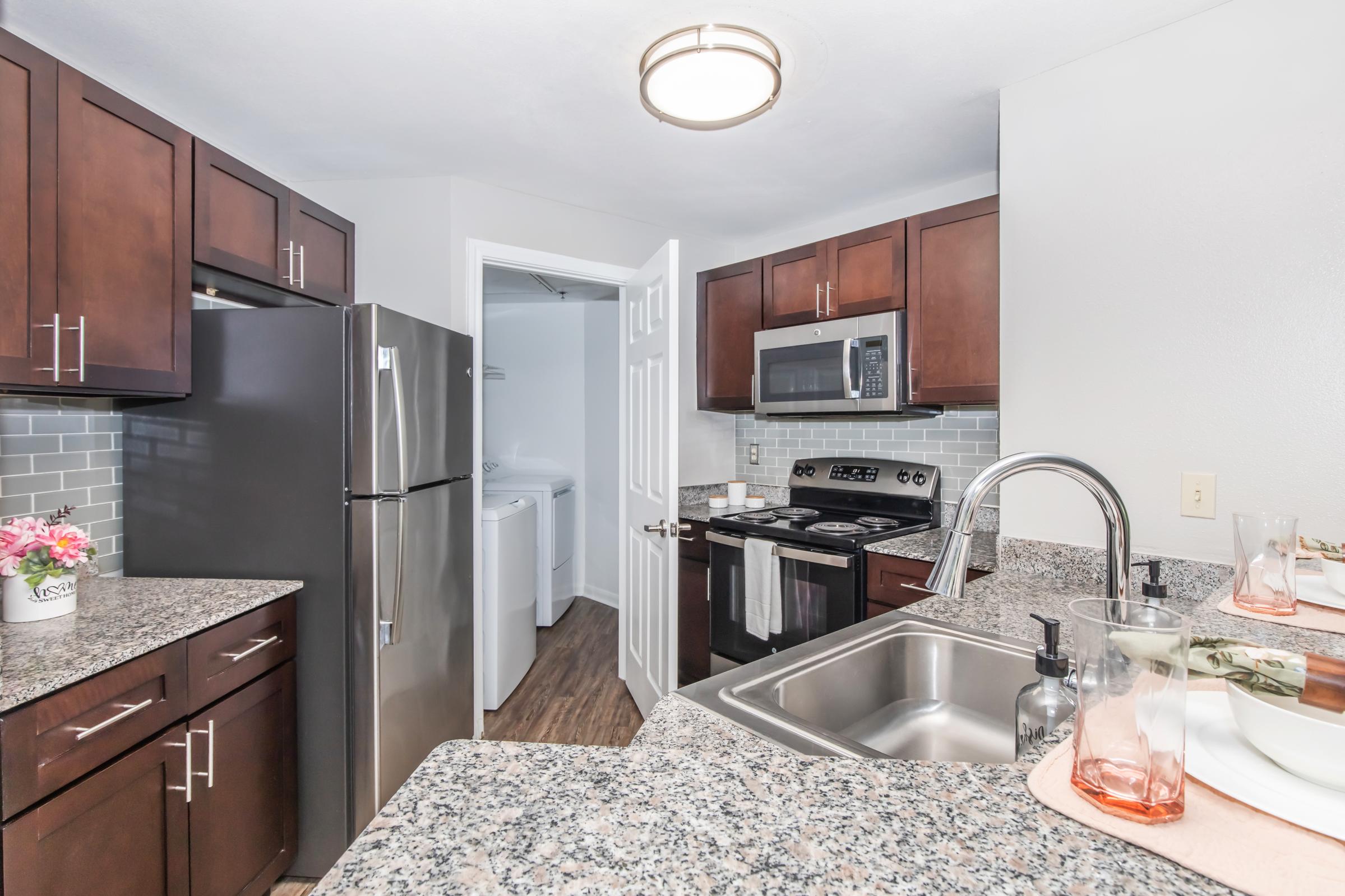 A modern kitchen featuring dark wood cabinets, stainless steel appliances, and granite countertops. The space includes a sink, stove, and refrigerator, with a view into a laundry area through an open door. Decorative items like a flower pot and glasses are placed on the counter, adding a cozy touch.