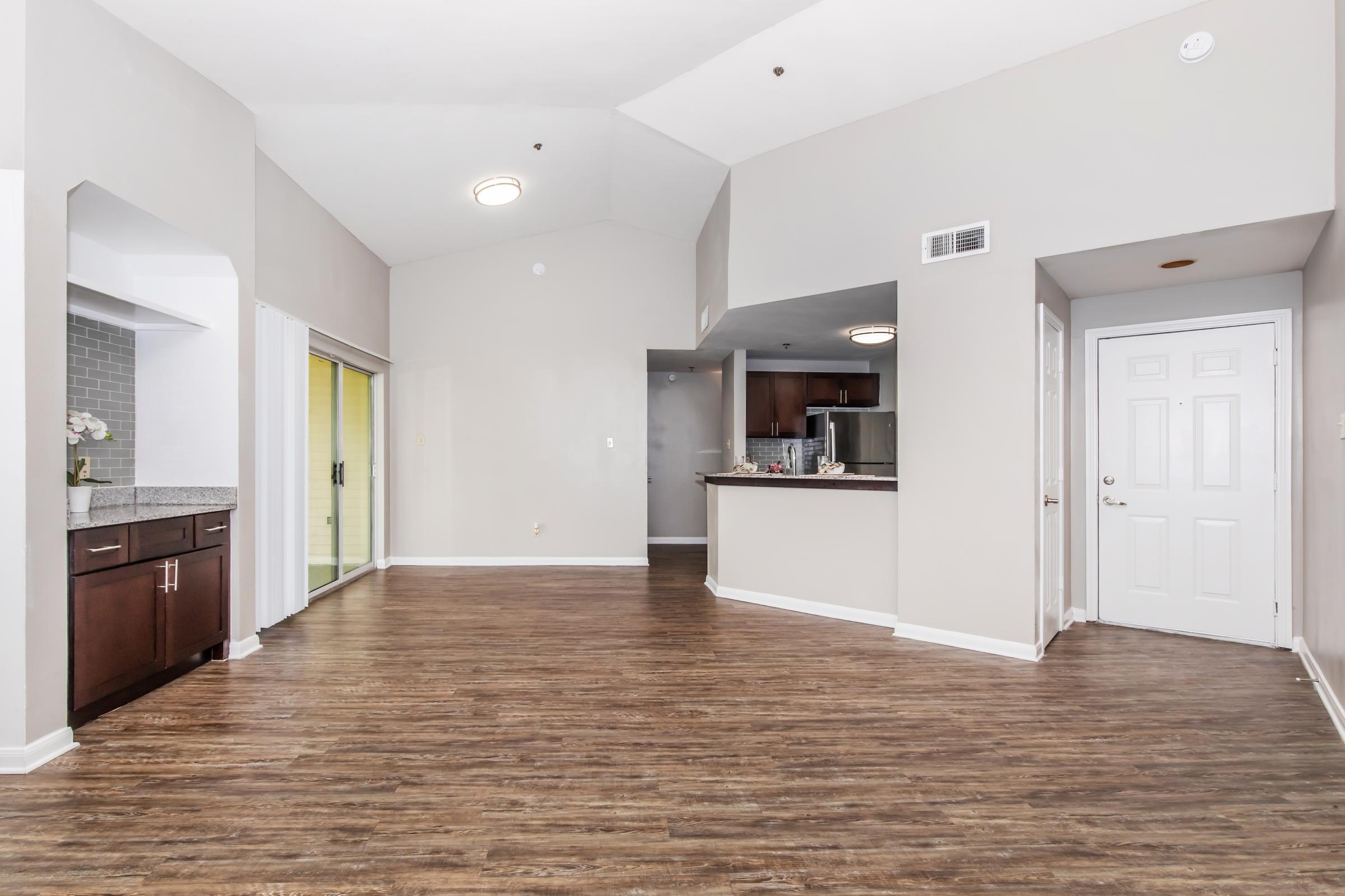 Spacious interior of an apartment featuring a living area with hardwood floors, arched doorway, and open layout. The kitchen is visible in the background with dark cabinets, and there are large windows. A front door is seen on the right side. Natural light brightens the room, enhancing the modern ambiance.