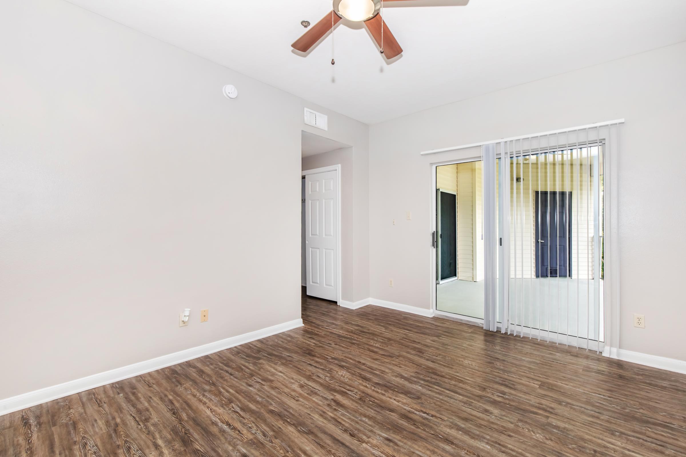 Interior view of a light-colored room featuring a ceiling fan and laminate wood flooring. A doorway leads to another room, and there is a sliding glass door that opens to an outdoor area. The walls are painted in neutral tones, creating a bright and spacious ambiance.